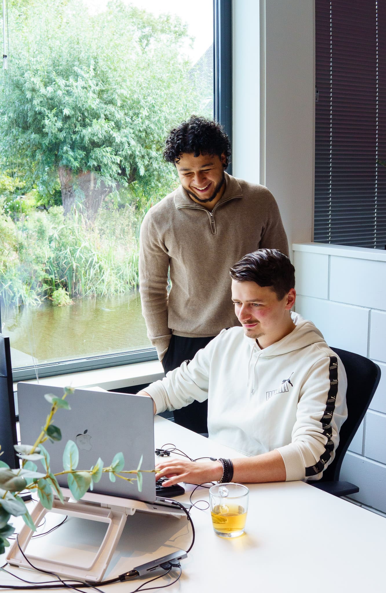 Twee personen werken samen aan een bureau bij een groot raam terwijl een van hen achter een laptop zit en de ander meekijkt.
