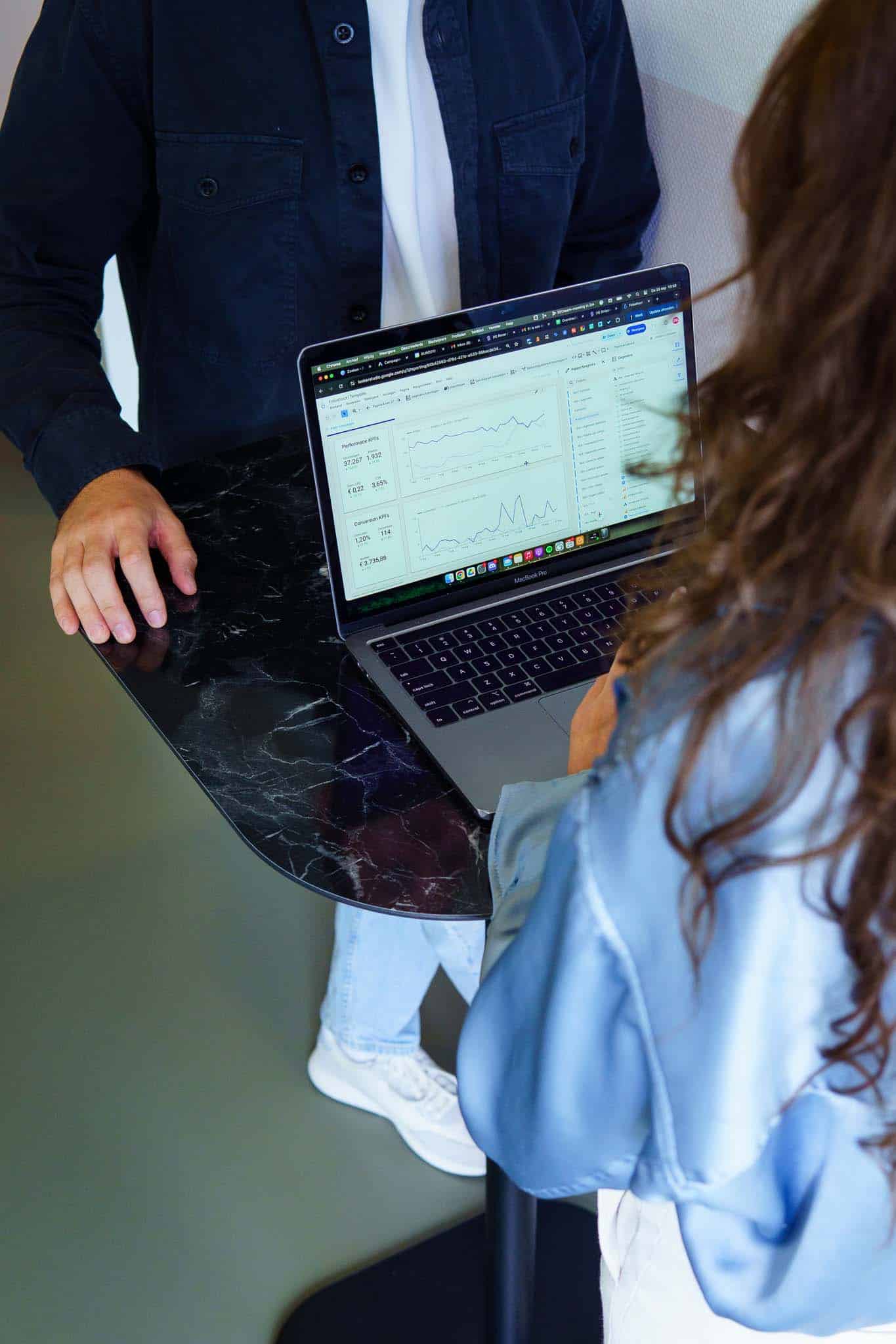 Twee personen staan bij een hoge tafel terwijl een laptop met grafieken en statistieken zichtbaar openstaat.