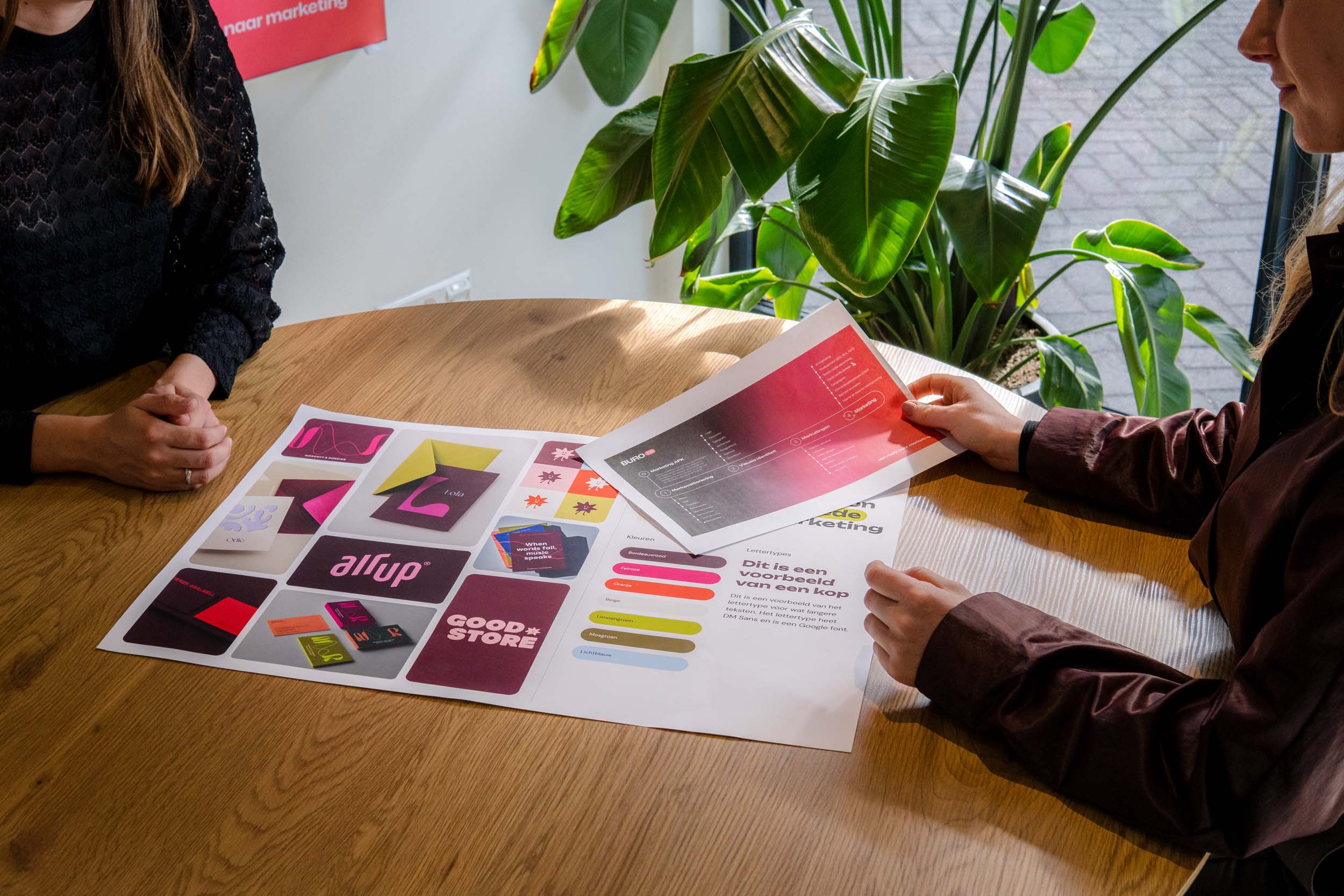 Twee personen bekijken drukwerk met brandingvoorbeelden en kleurstalen op een ronde houten tafel.