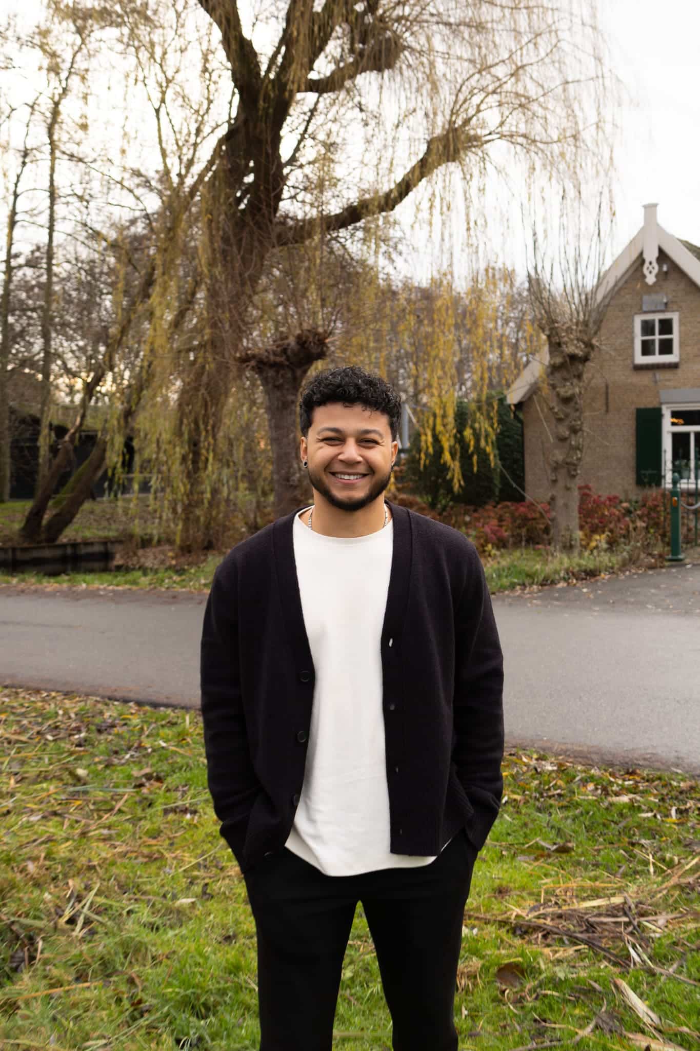 BURO210 Fullservice Marketingbureau Jonge, lachende man met krullend haar en casual kleding staat buiten voor een pittoresk Nederlands huis met een wilg. Mooie herfstachtige achtergrond.