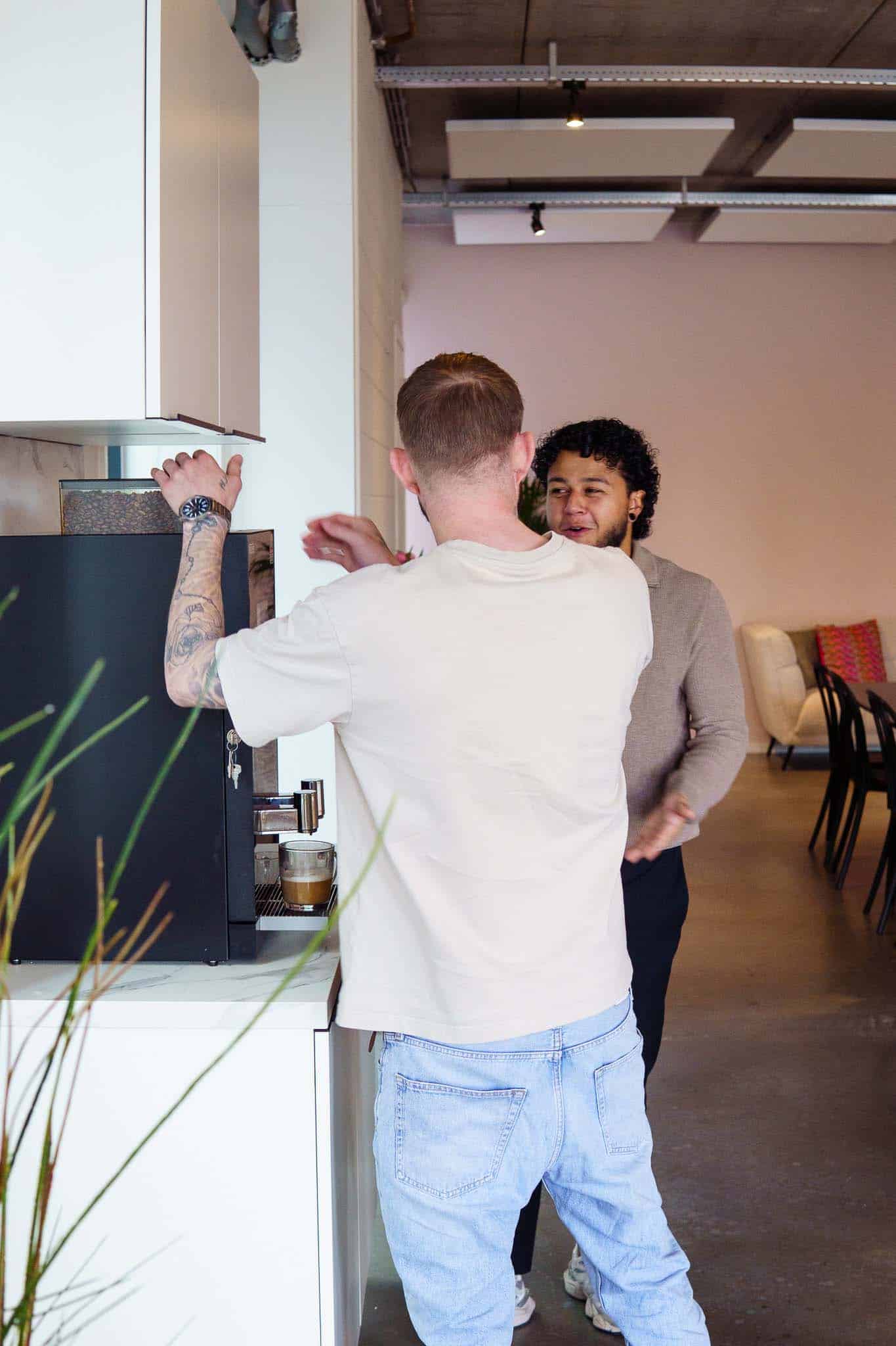 BURO210 Fullservice Marketingbureau Twee mannen in gesprek bij een moderne koffiemachine. Een collega bereidt koffie in een stijlvol kantoor, terwijl de andere toekijkt. Interieur met design meubels.