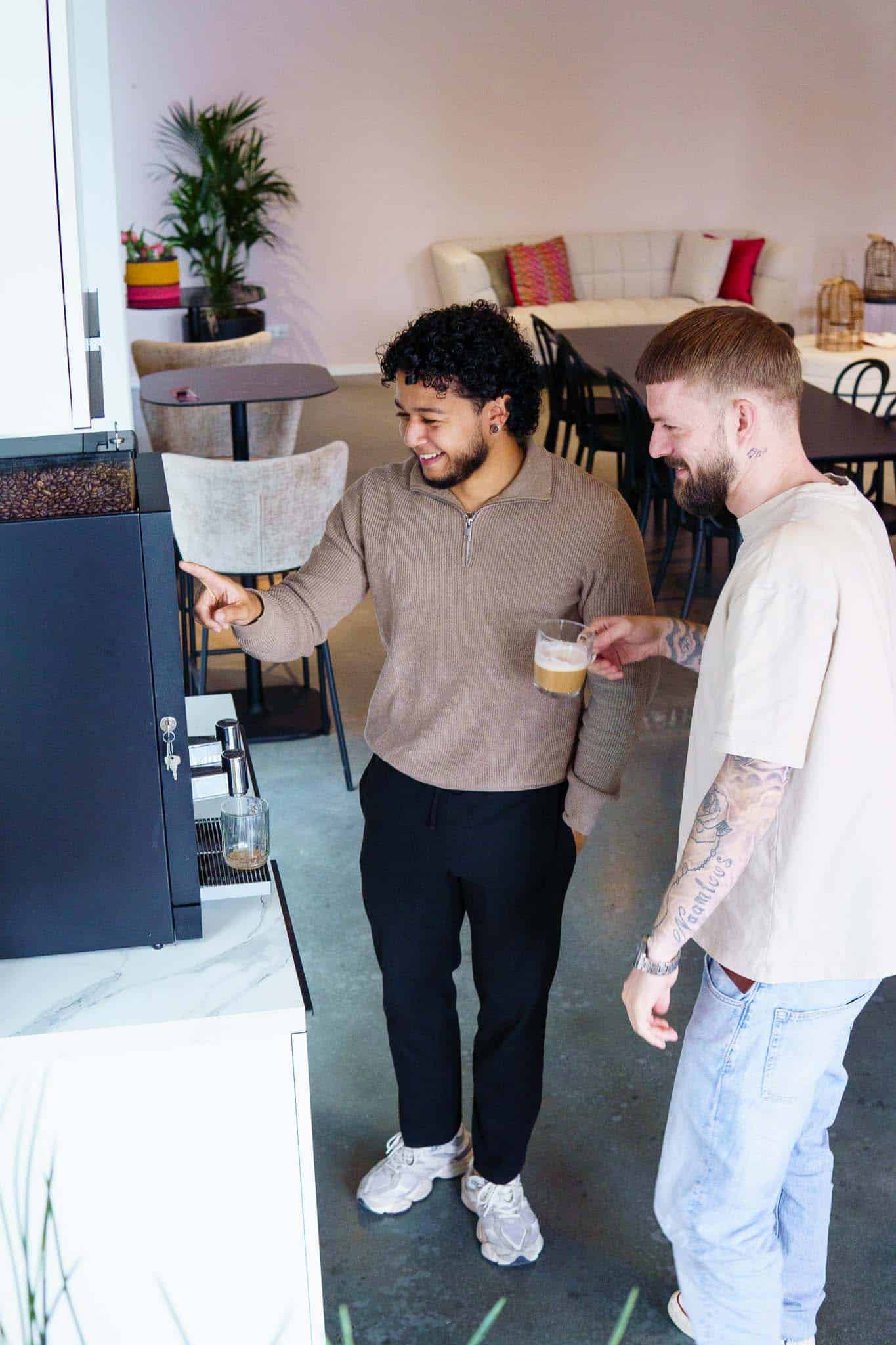 BURO210 Fullservice Marketingbureau Twee mannen lachen bij een koffiemachine in een moderne kantoorruimte. Eén wijst naar de machine, terwijl de ander een glas koffie vasthoudt, omringd door stijlvol meubilair.