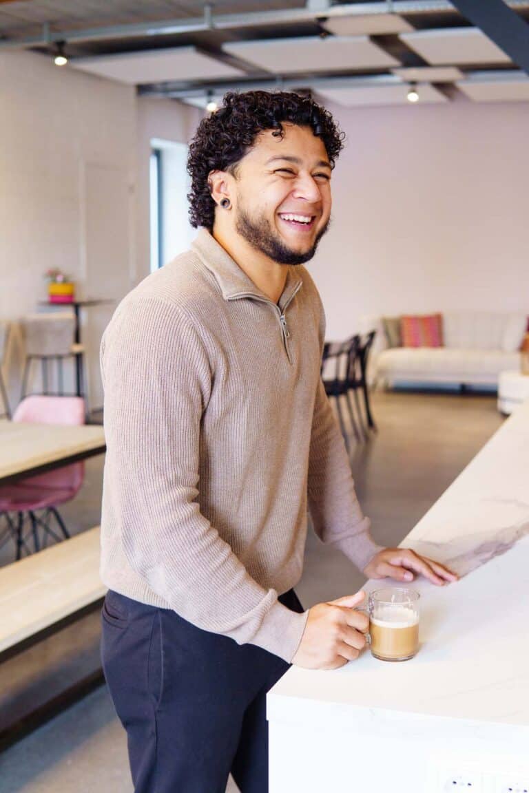BURO210 Fullservice Marketingbureau Lachende jonge man met krullend haar geniet van een latte in een moderne kantoorruimte. Hij staat ontspannen bij een marmeren aanrecht.