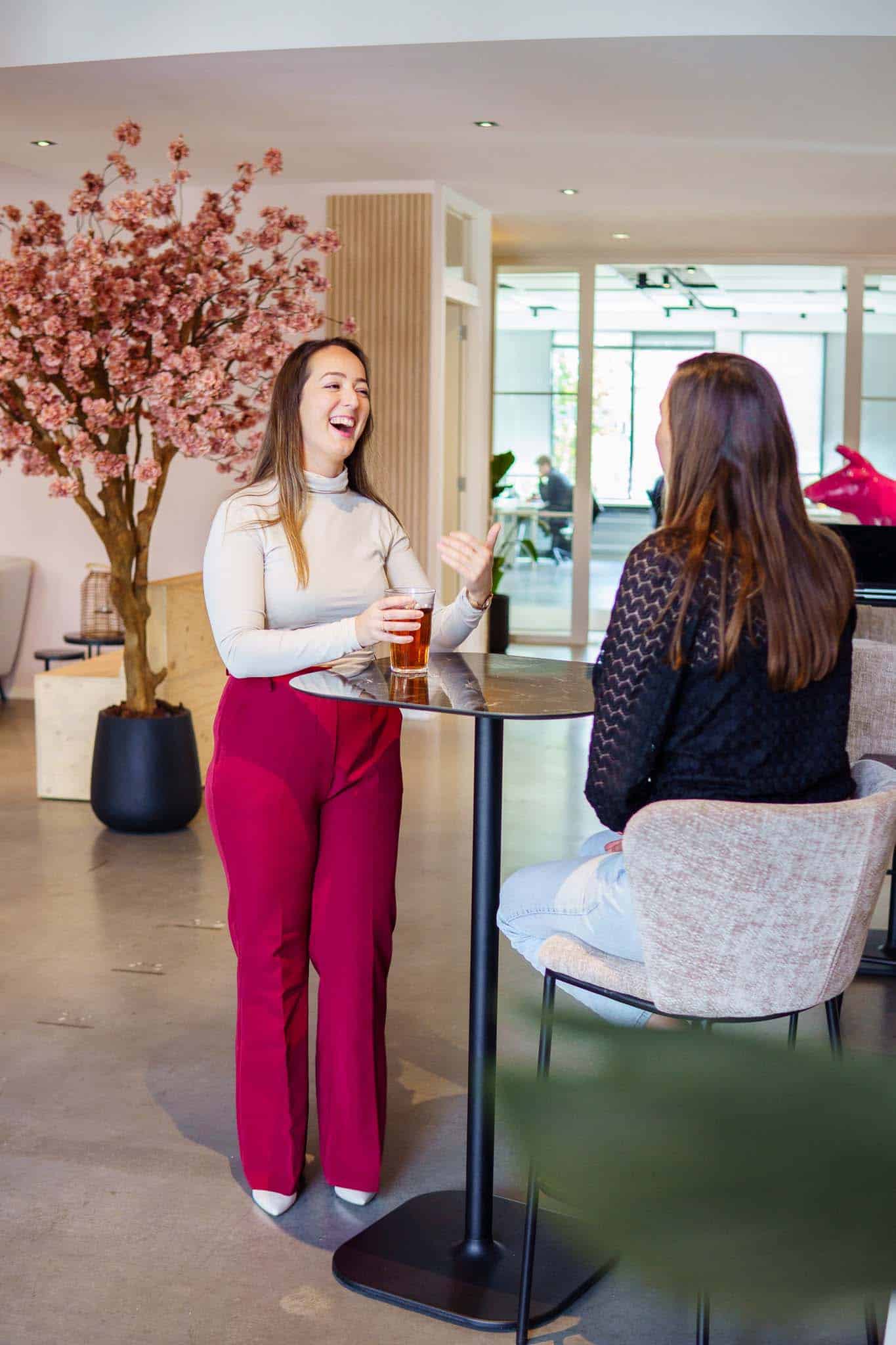 BURO210 Fullservice Marketingbureau Twee collega's hebben een informeel gesprek tijdens een pauze in een modern kantoor, met een drankje aan een statafel en een decoratieve bloesemboom.