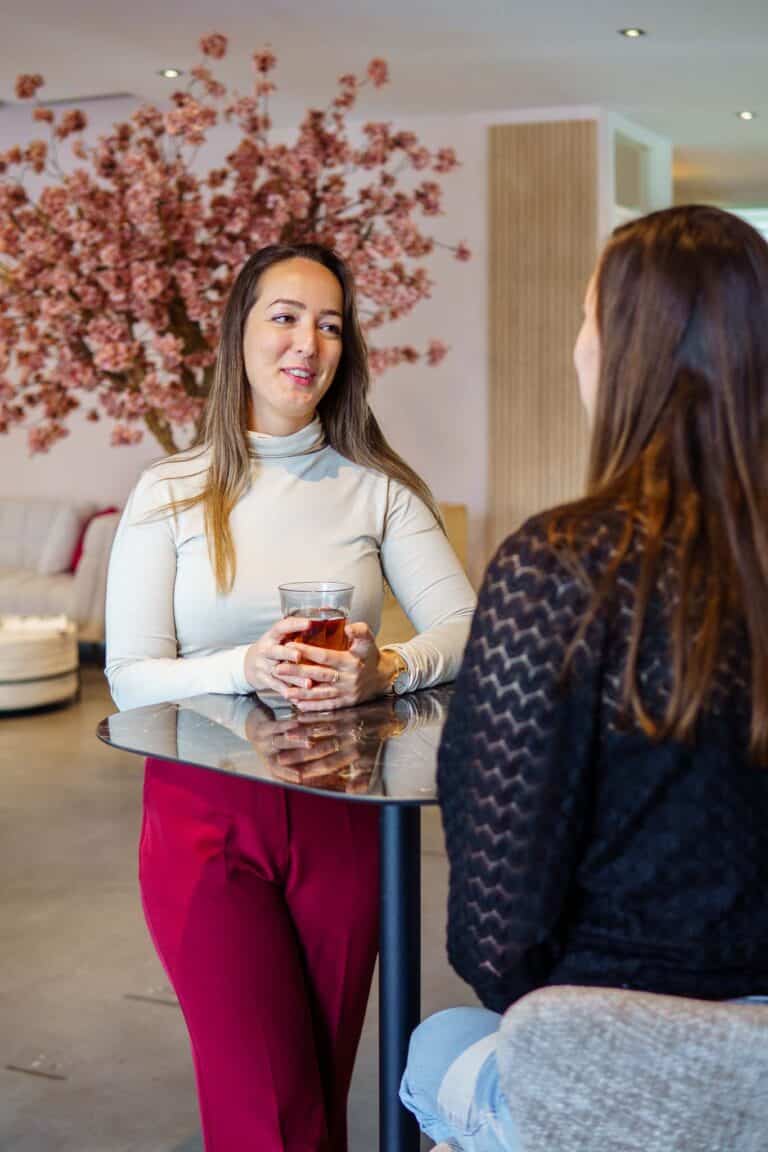 Twee personen staan bij een hoge tafel in een kantoorruimte en praten terwijl een van hen een glas thee vasthoudt.