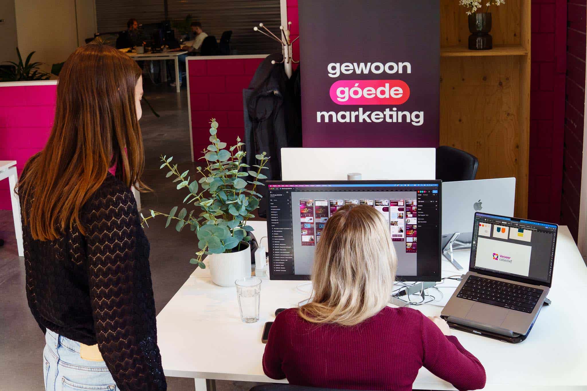 Twee personen werken aan een bureau met een groot beeldscherm en laptop waarop marketingontwerpen te zien zijn.