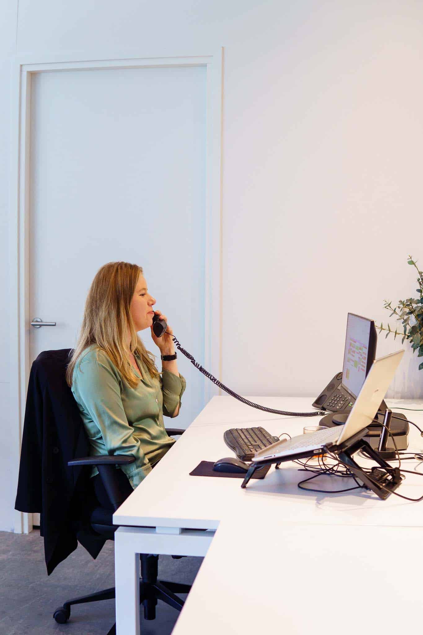 BURO210 Fullservice Marketingbureau Professionele vrouw aan het werk op kantoor, telefonerend met een klassieke telefoonhoorn. Laptop en computerscherm zichtbaar op een wit bureau.