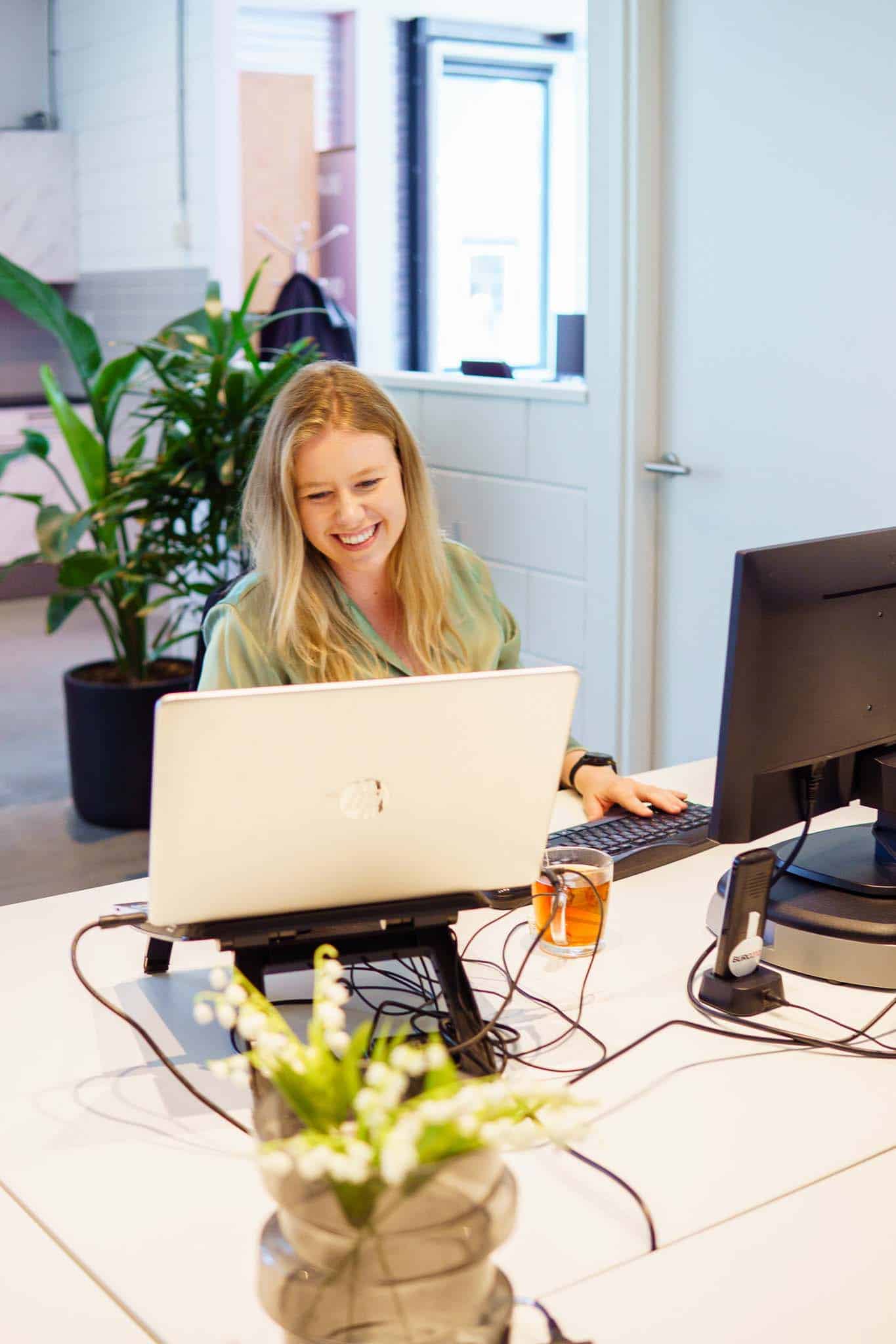 BURO210 Fullservice Marketingbureau Vrolijke jonge vrouw werkt op een laptop aan een bureau op kantoor. Ze lacht en typt op het toetsenbord, met een kop thee naast de computer.