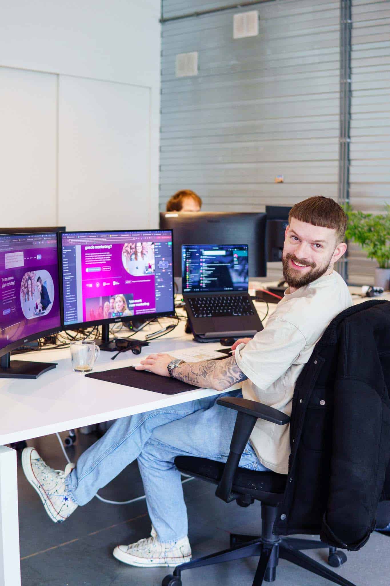 BURO210 Fullservice Marketingbureau Vrolijke jonge man met baard zit aan een bureau met computerschermen in een modern kantoor. Hij glimlacht en kijkt over zijn schouder.