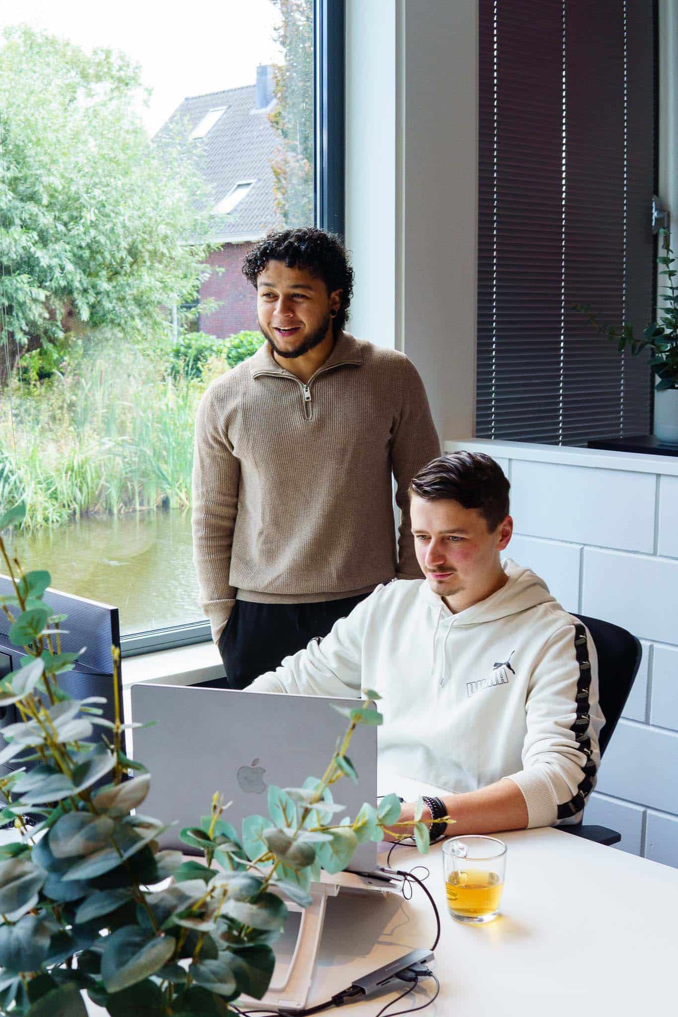 BURO210 Fullservice Marketingbureau Twee jonge mannen werken samen in een modern kantoor met uitzicht op het water. Eén persoon zit aan een laptop, de ander kijkt uit het raam.