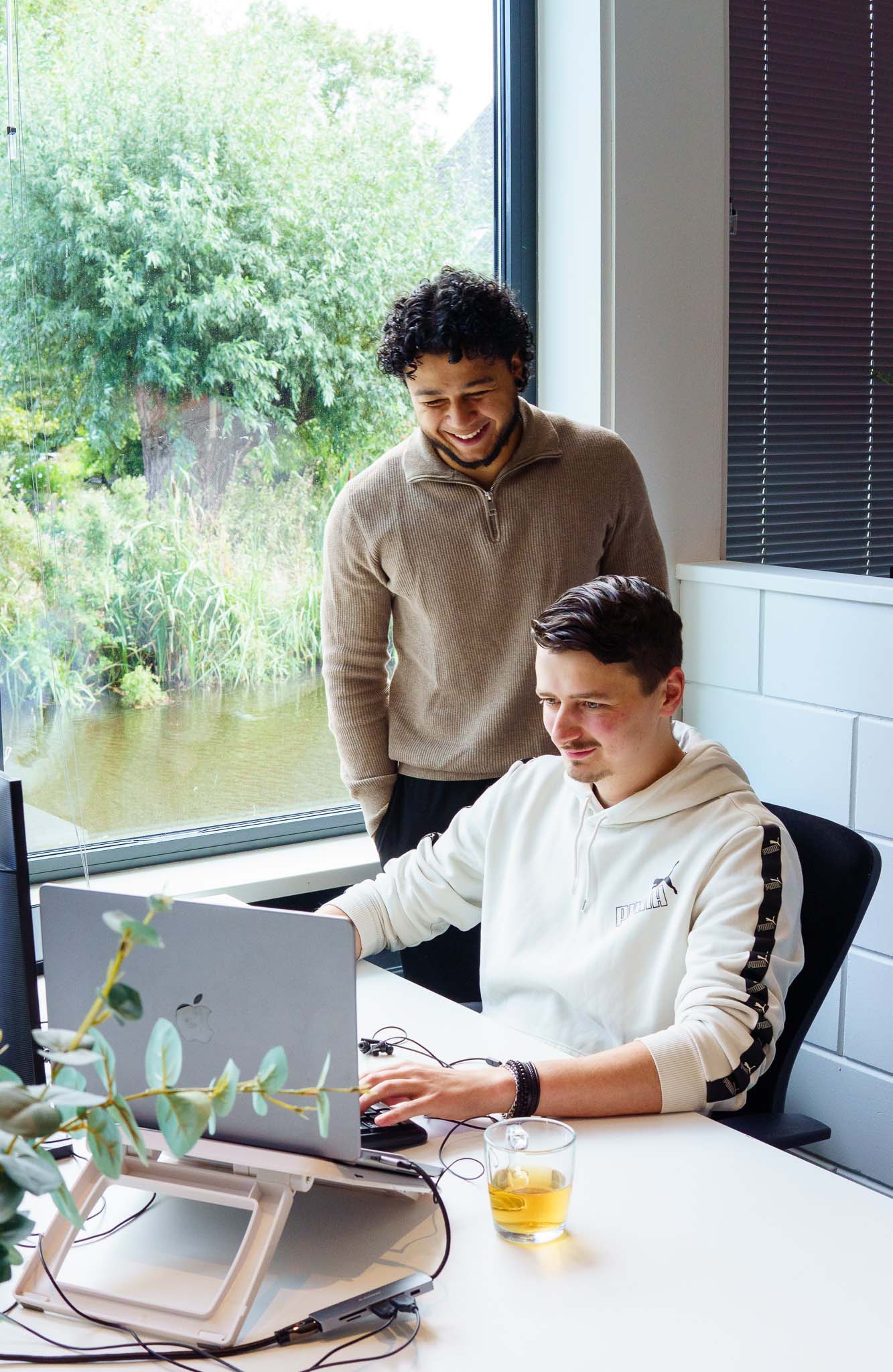 BURO210 Fullservice Marketingbureau Twee collega's werken samen aan een laptop in een modern kantoor met uitzicht op een rivier. De ene persoon zit, de ander staat en kijkt toe.