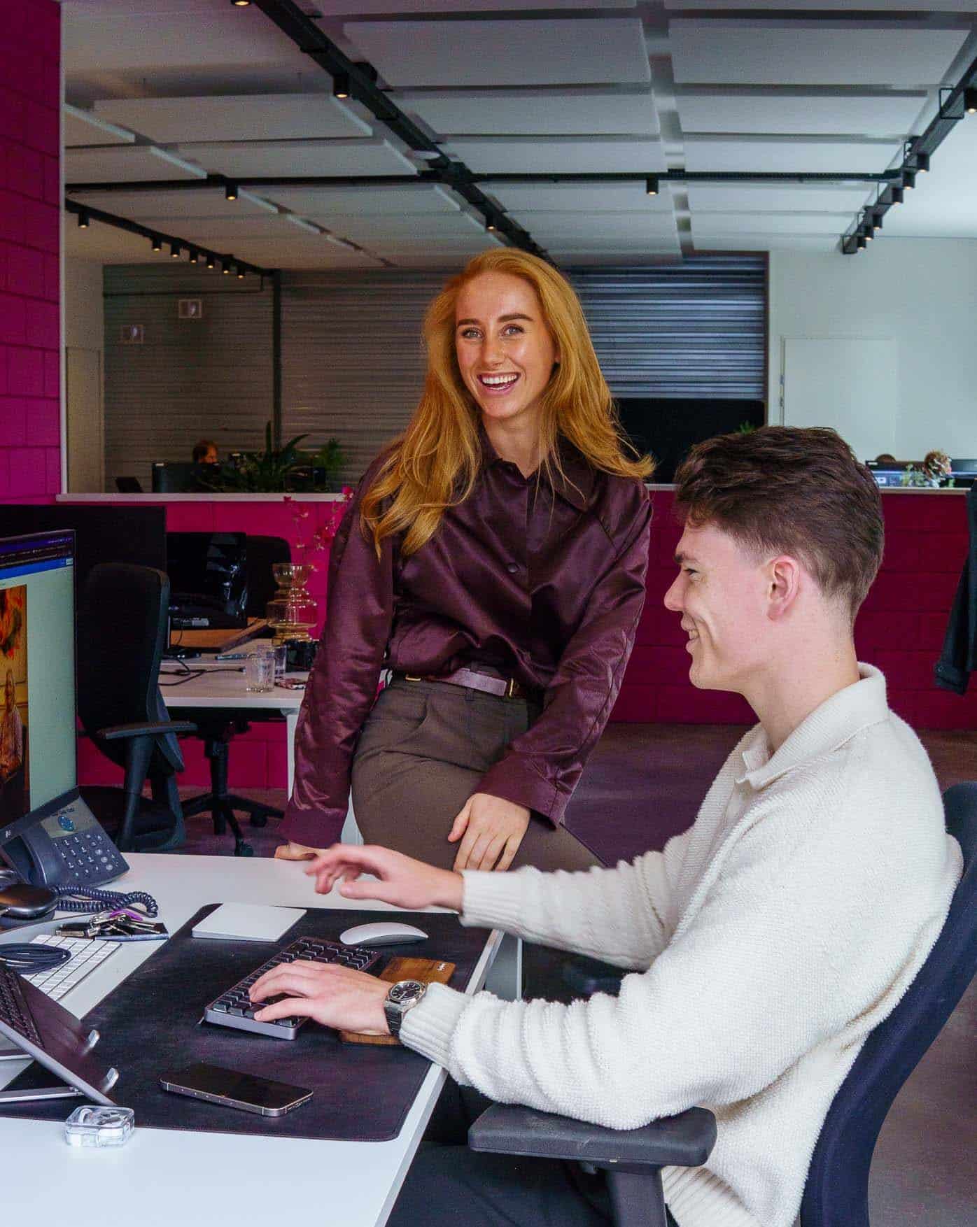 BURO210 Fullservice Marketingbureau Twee jonge collega's in een modern kantoor: een man typt op een toetsenbord terwijl een vrouw met lang rood haar lachend toekijkt. Samenwerking en teamwerk.