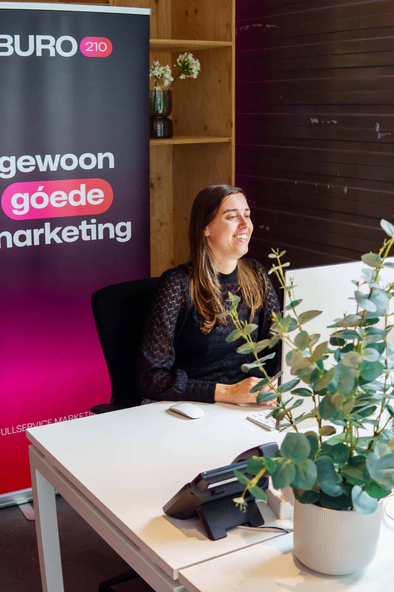 BURO210 Fullservice Marketingbureau Lachende vrouw achter bureau bij BURO 210, een fullservice marketing bureau, met bloemen en planten voor een productieve en aangename werkomgeving.