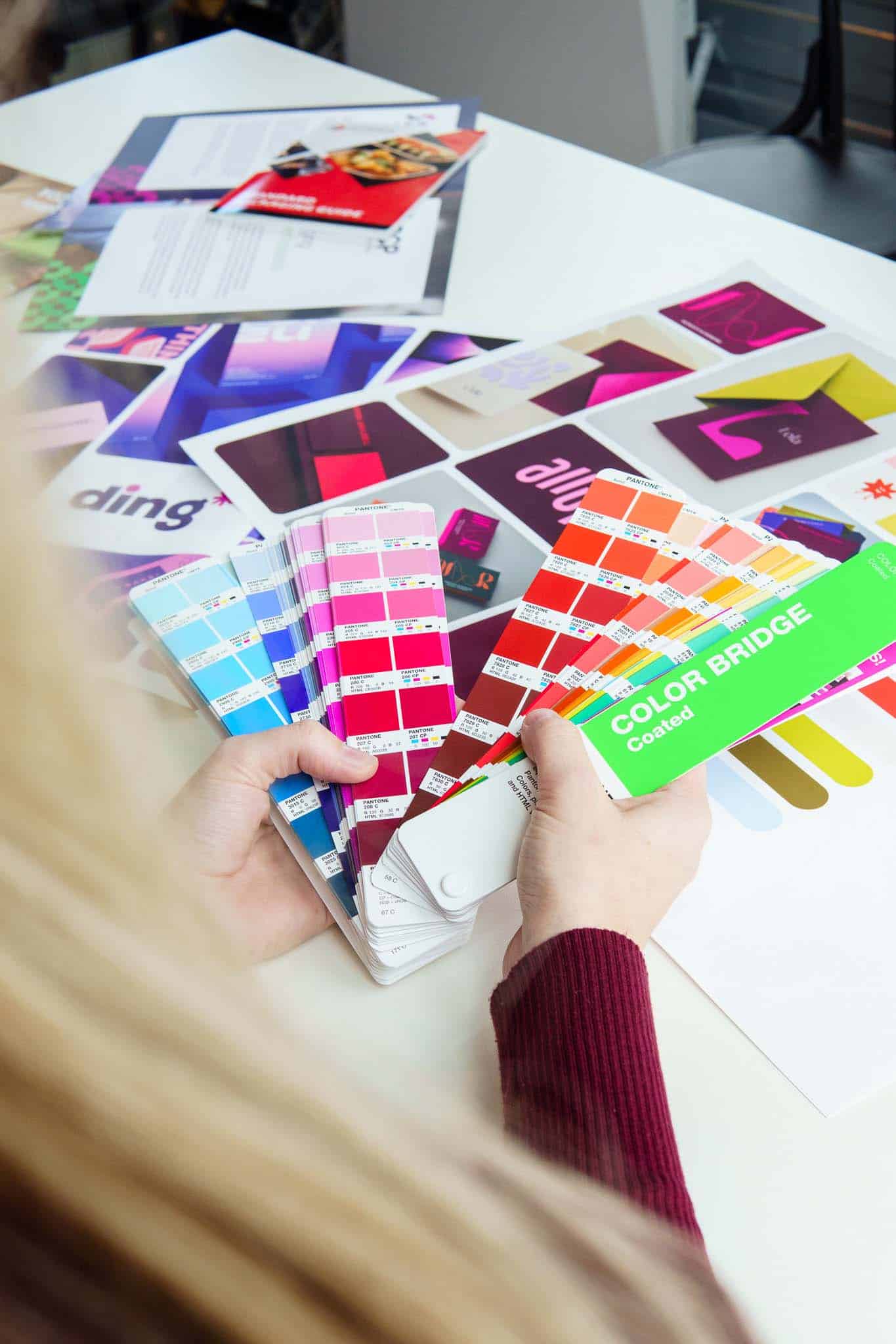 BURO210 Fullservice Marketingbureau Persoon houdt Pantone kleurstaalkaarten vast boven een witte tafel met kleurrijke ontwerpen. Kleurkeuze en grafisch ontwerp concept. Pantone Color Bridge coated voor inspiratie.