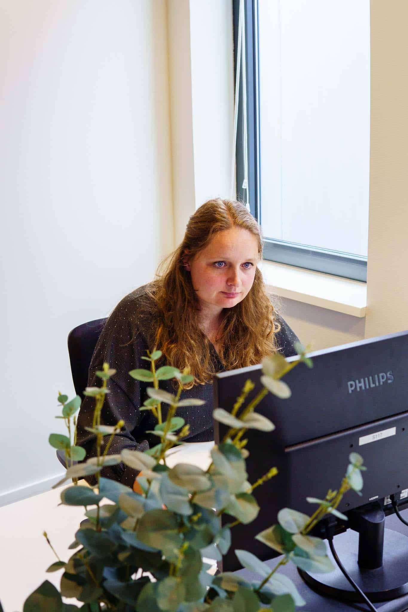 BURO210 Fullservice Marketingbureau Vrouw met bruin haar werkt geconcentreerd achter een Philips monitor in een lichte kantooromgeving met plant op het bureau.