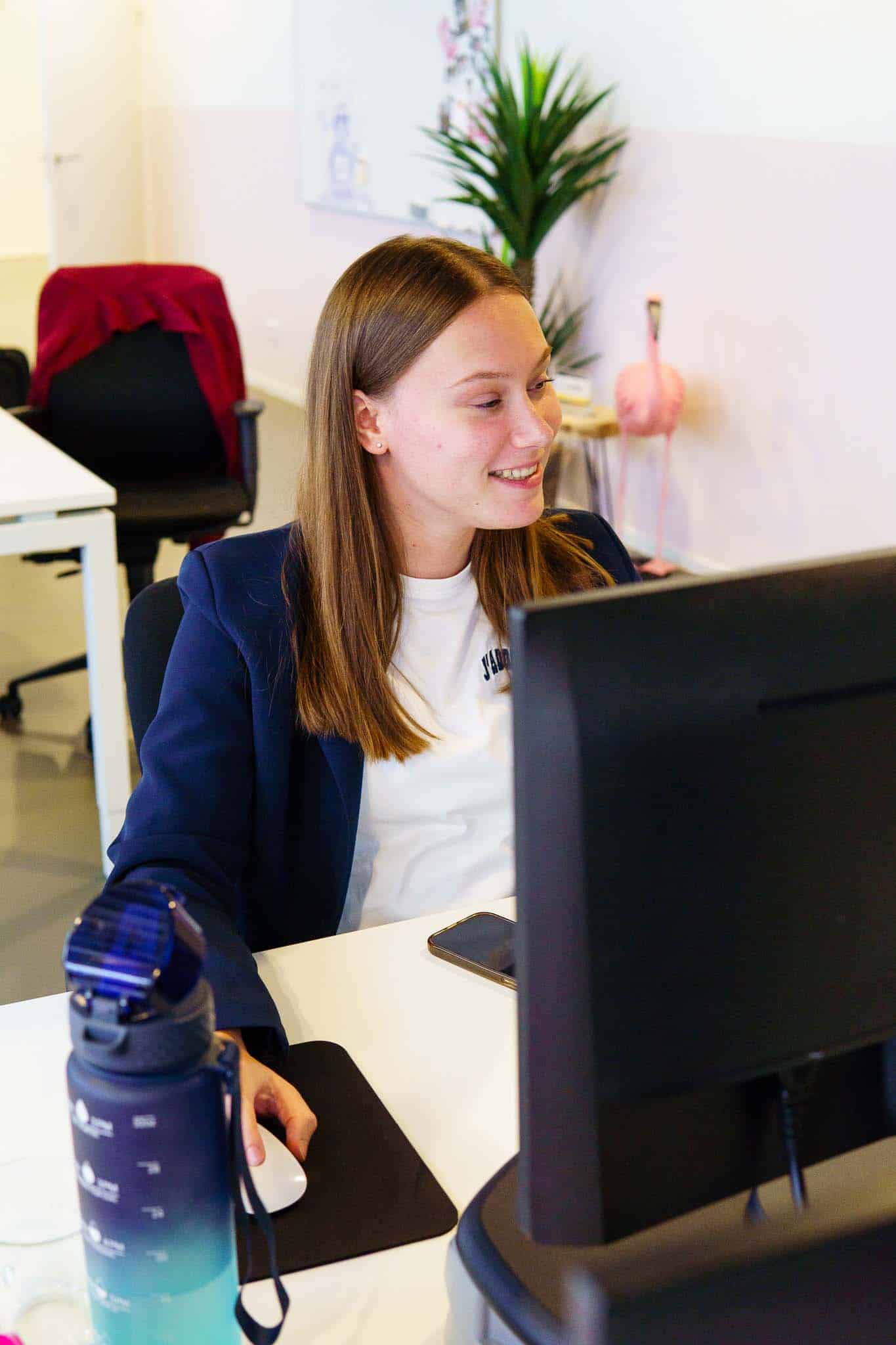 BURO210 Fullservice Marketingbureau Jonge professionele vrouw lacht tijdens het werken op een computer in een modern kantoor, met een waterfles en telefoon op het bureau.