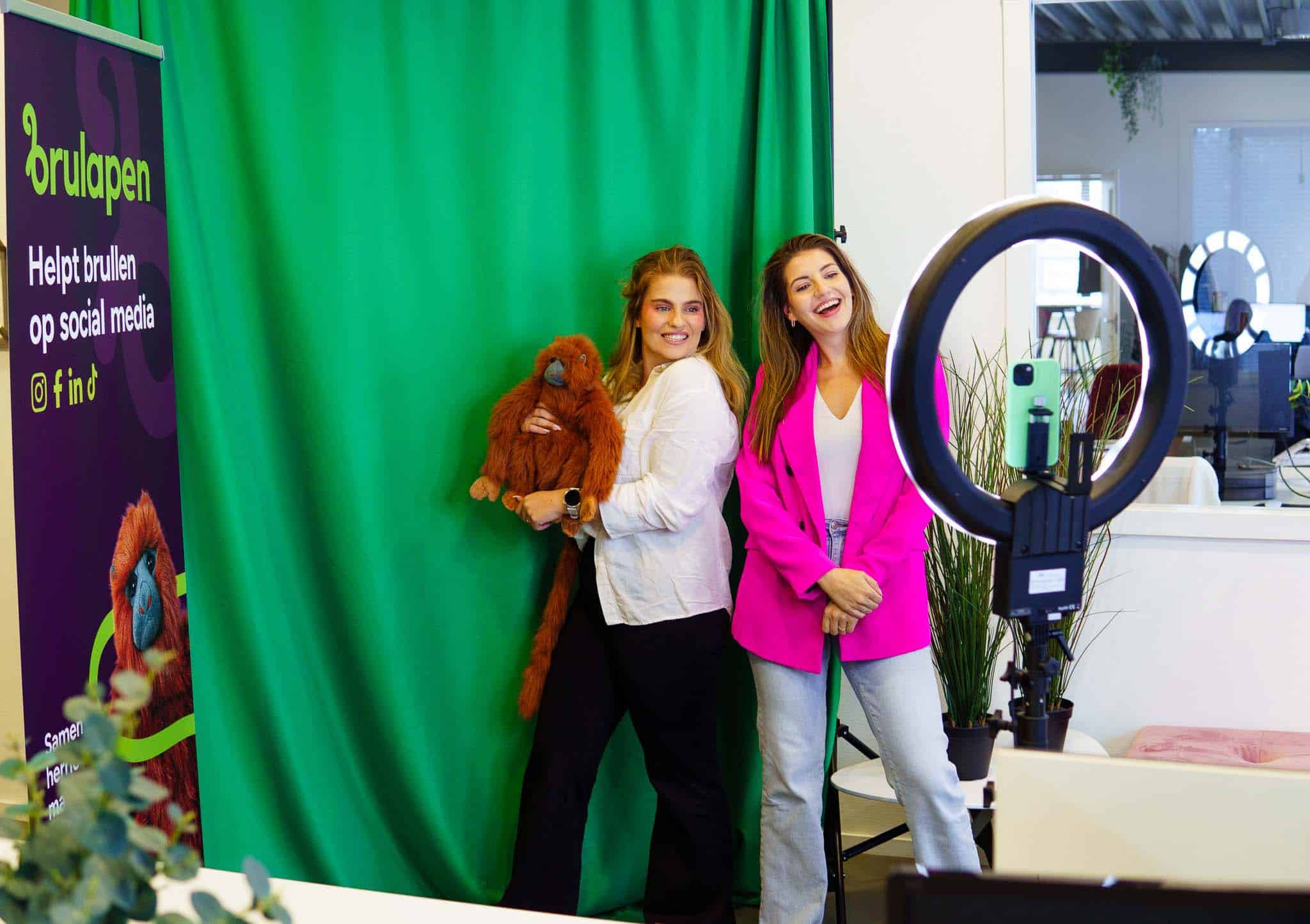 BURO210 Fullservice Marketingbureau Twee vrolijke vrouwen poseren met een knuffelaap voor een groene achtergrond, gefotografeerd met een ringlamp voor sociale media content.