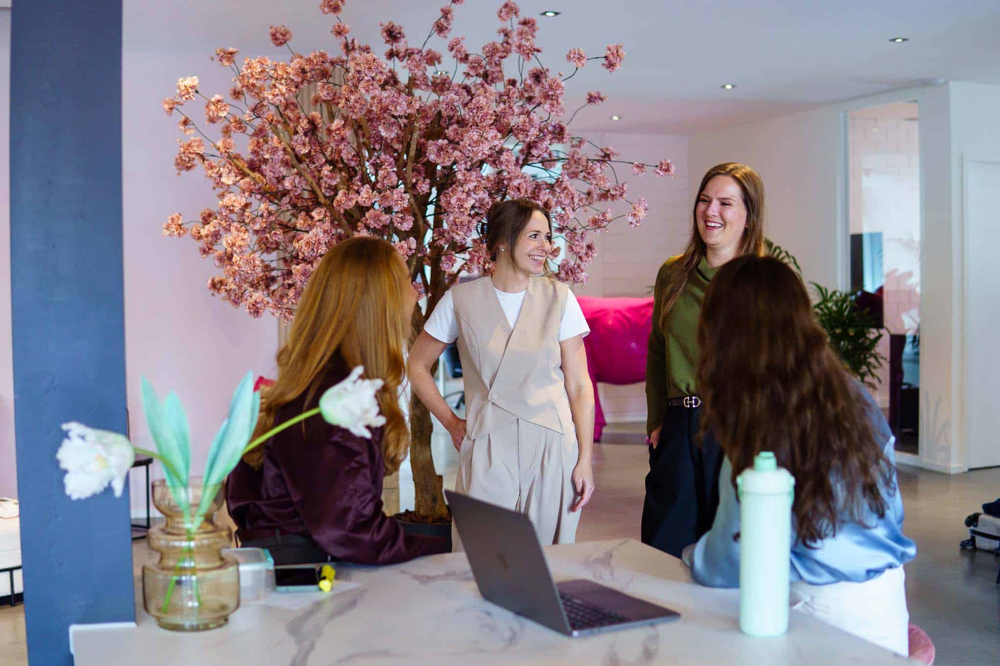 BURO210 Fullservice Marketingbureau Een groep van vier vrolijke vrouwen in een lichte, moderne kantoorruimte, in gesprek met een laptop op een tafel en een bloeiende kersenboom op de achtergrond.