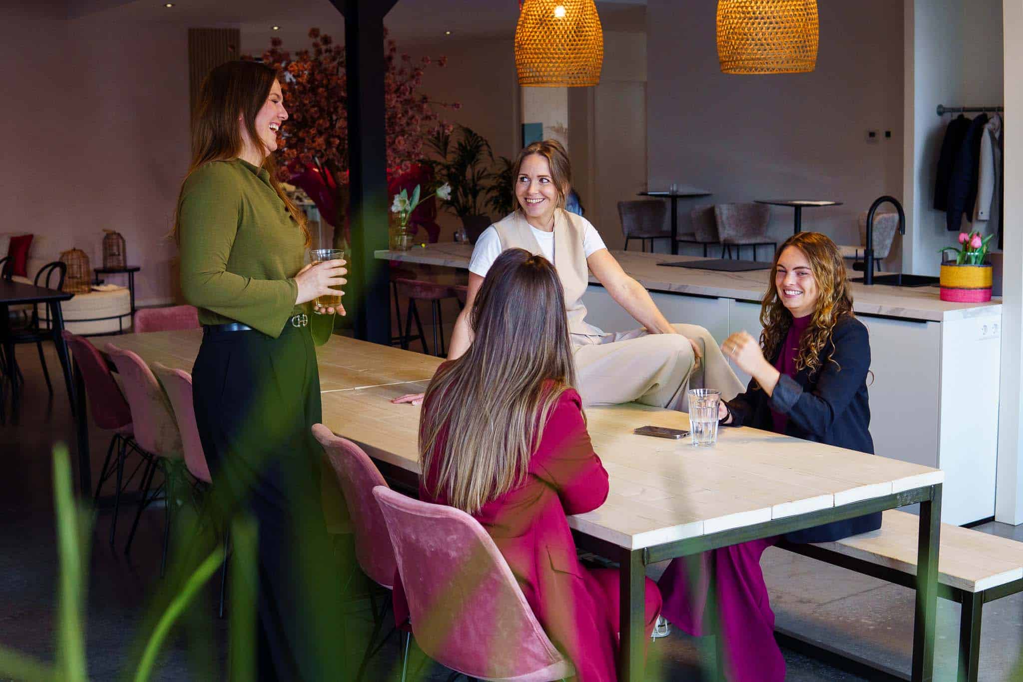 BURO210 Fullservice Marketingbureau Groep van vier lachende vrouwen verzameld rond een grote houten tafel in een lichte, moderne ruimte. Ze lijken een gesprek te voeren en genieten van elkaars gezelschap.