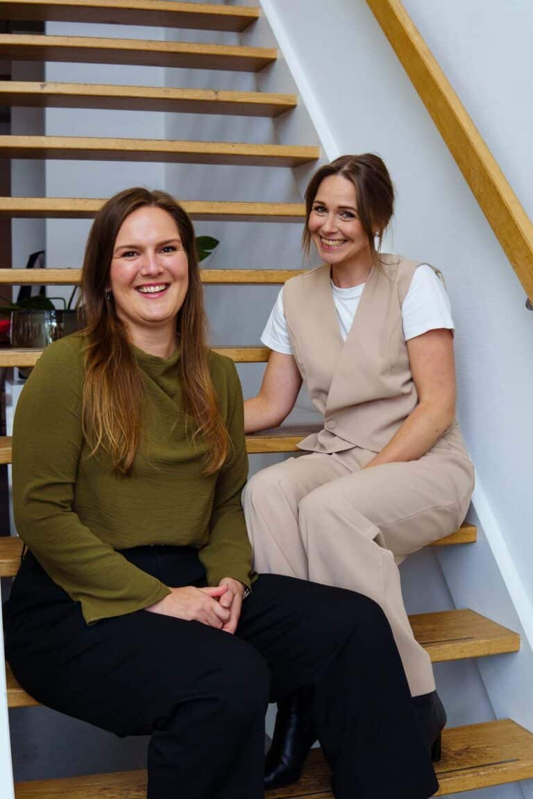 BURO210 Fullservice Marketingbureau Twee lachende vrouwen zitten op een houten trap in een huiselijke setting. De vrouwen dragen stijlvolle kleding en poseren voor een foto in een heldere, natuurlijke omgeving.