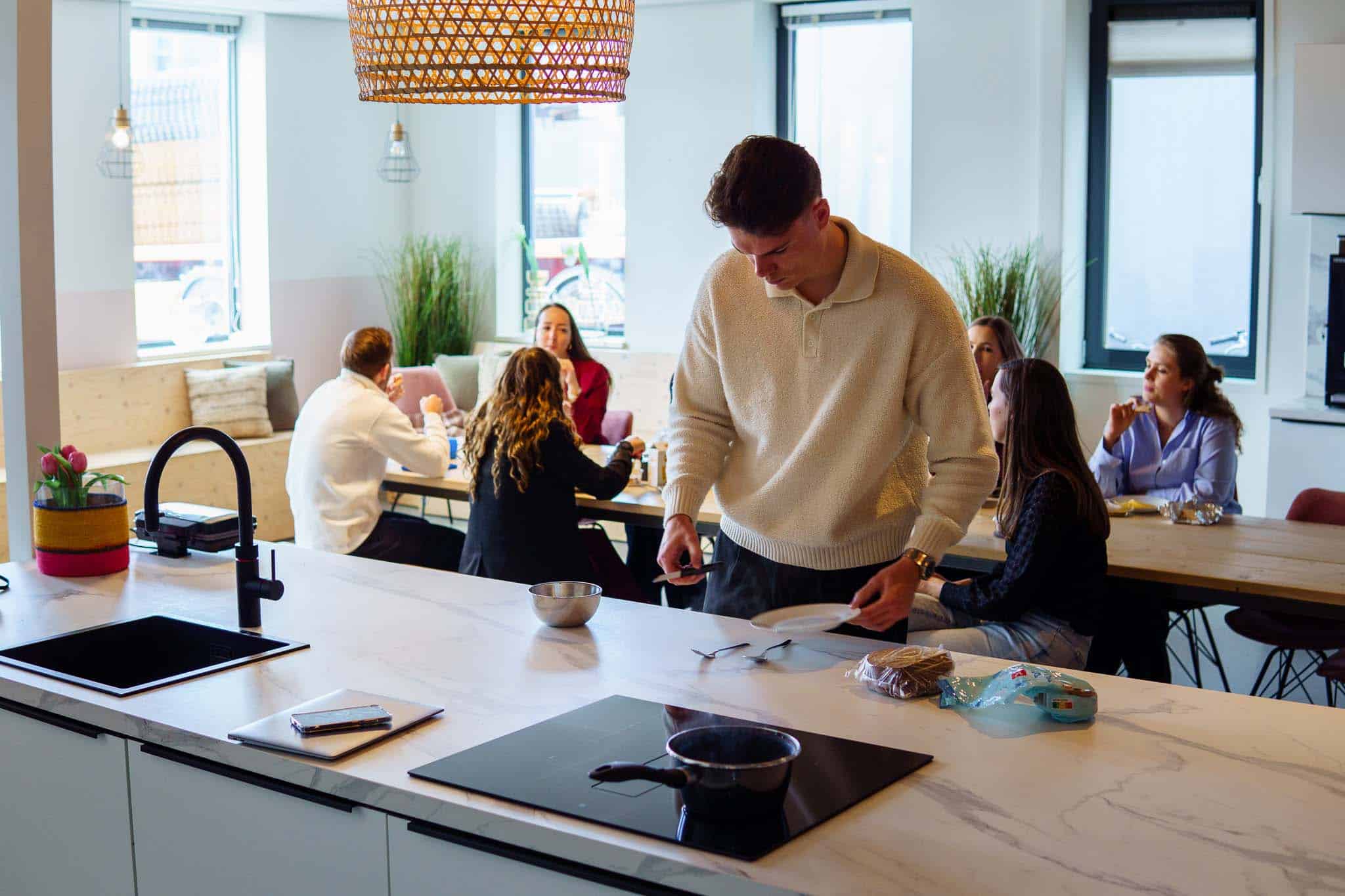 BURO210 Fullservice Marketingbureau Moderne gedeelde keukenruimte met een jongeman die borden klaarmaakt voor een maaltijd met vrienden. Een gezellige en informele sfeer met natuurlijk licht en design verlichting.