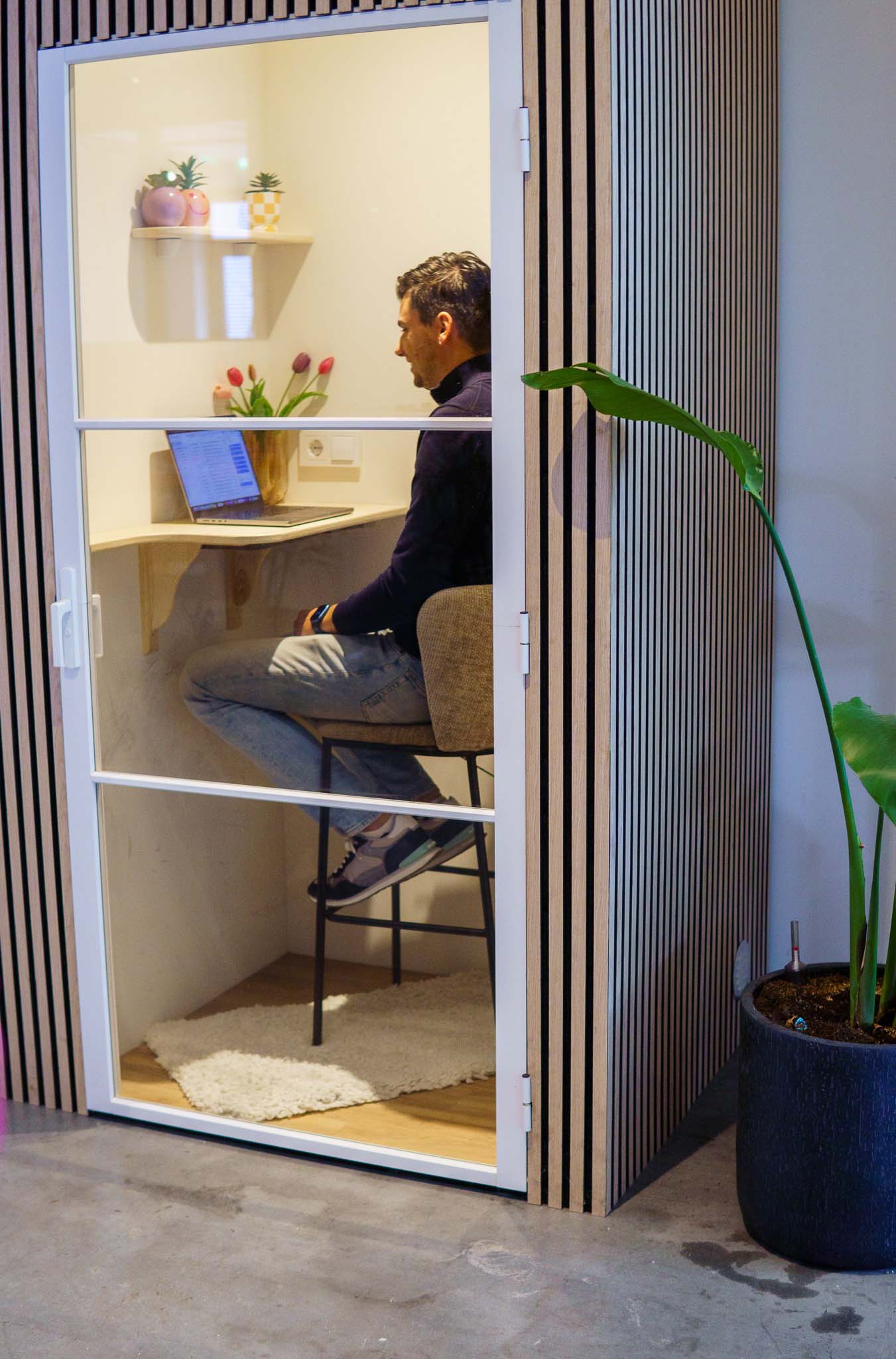 BURO210 Fullservice Marketingbureau Een man werkt geconcentreerd op zijn laptop in een geluidsdichte kantoorruimte. De cabine heeft een modern design met houten lambrisering en een glazen wand.