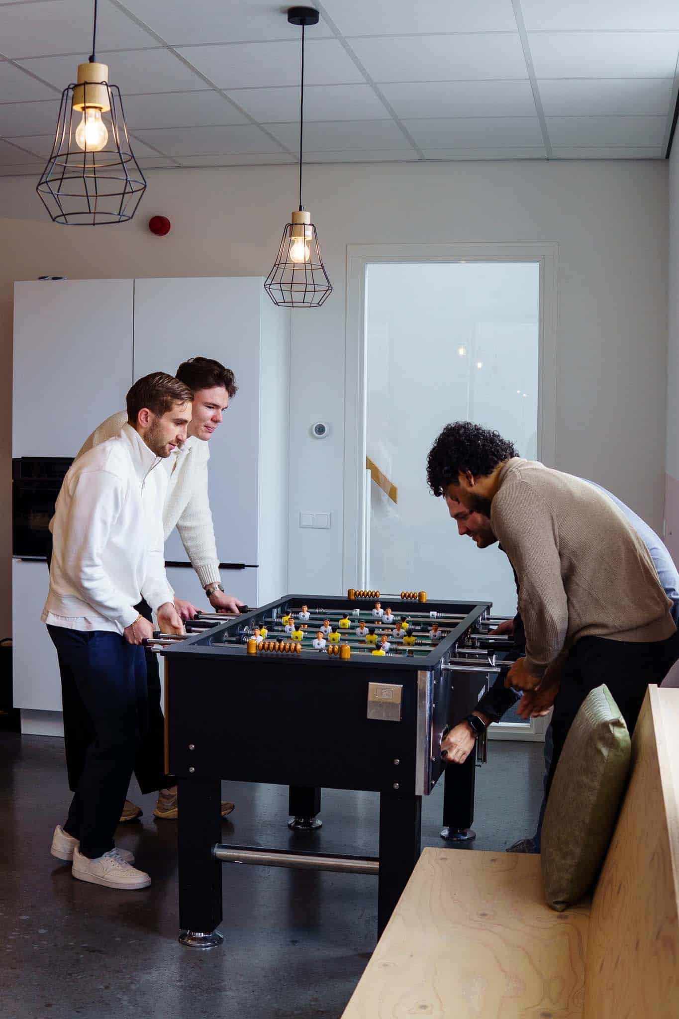 BURO210 Fullservice Marketingbureau Drie mannen spelen tafelvoetbal in een lichte, moderne kantooromgeving. De focus ligt op plezier en teambuilding tijdens een pauze.