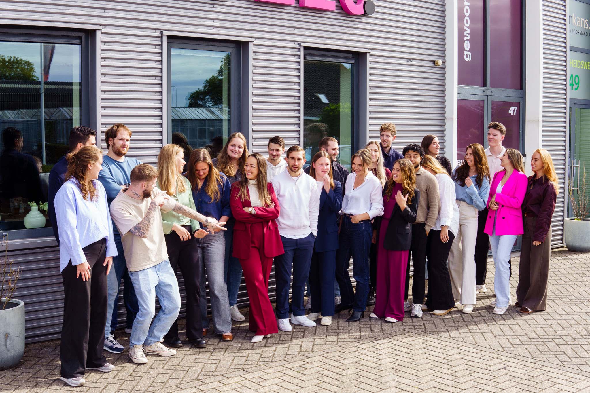 BURO210 Fullservice Marketingbureau Groepsfoto van een divers team voor het kantoorgebouw van Gewoon Makelaars. De medewerkers staan glimlachend op de foto, gekleed in kleurrijke kleding.