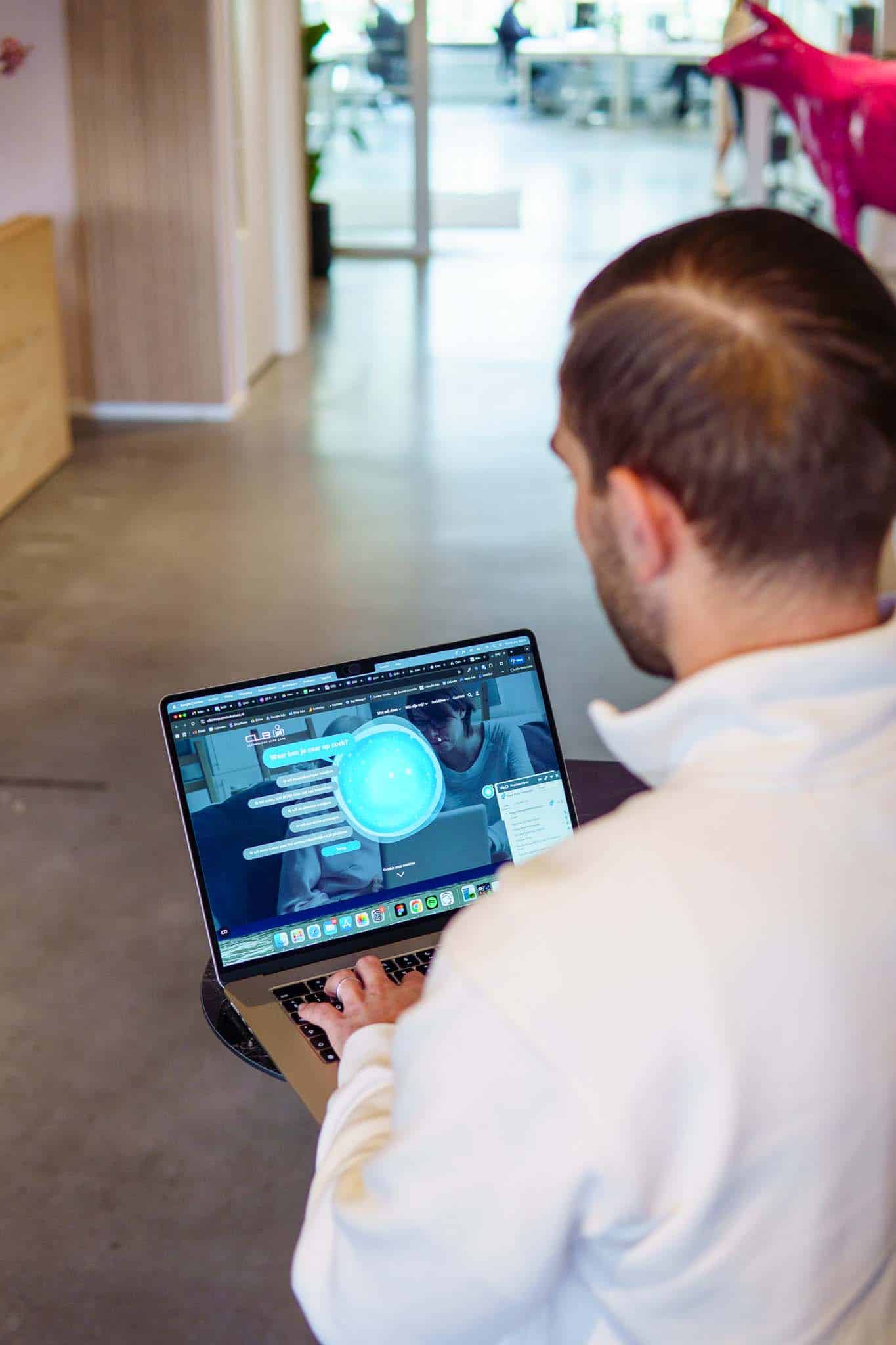 BURO210 Fullservice Marketingbureau Man kijkt naar laptop in een moderne kantooromgeving. Het laptopscherm toont een website met de tekst 'Waar ben je naar op zoek?'. Achtergrond bevat een roze sculptuur.