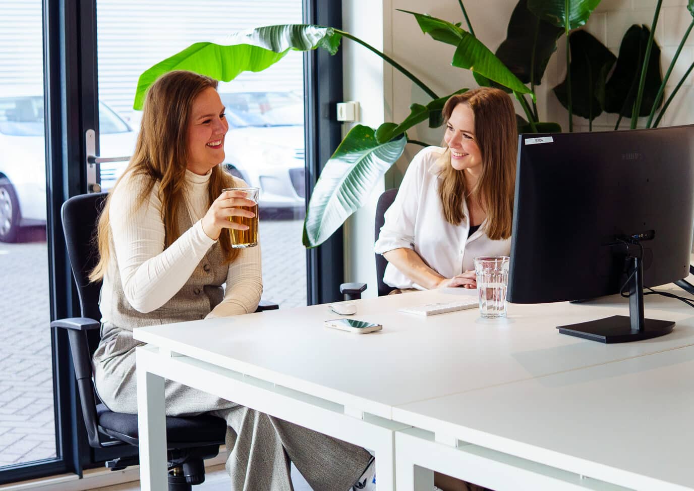 BURO210 Fullservice Marketingbureau Twee jonge, lachende vrouwen aan een bureau in een modern kantoor met planten en computers. Relaxte sfeer tijdens de werkdag.