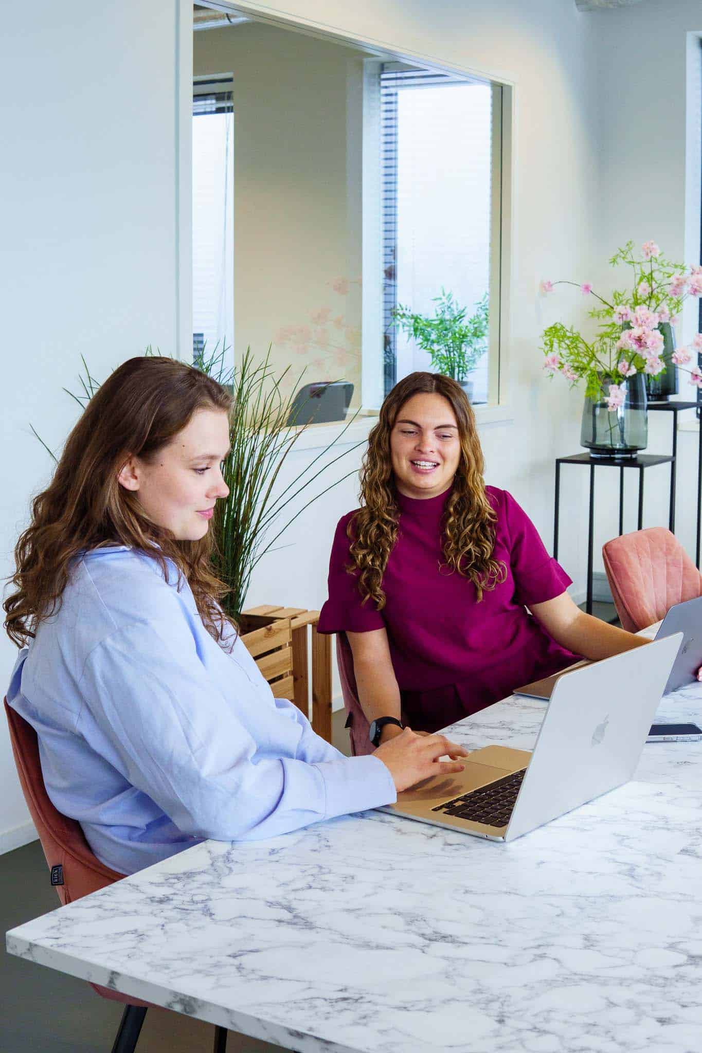 BURO210 Fullservice Marketingbureau Twee jonge, lachende vrouwen werken samen aan een laptop in een lichte en moderne kantoorruimte met planten en bloemen op tafel.