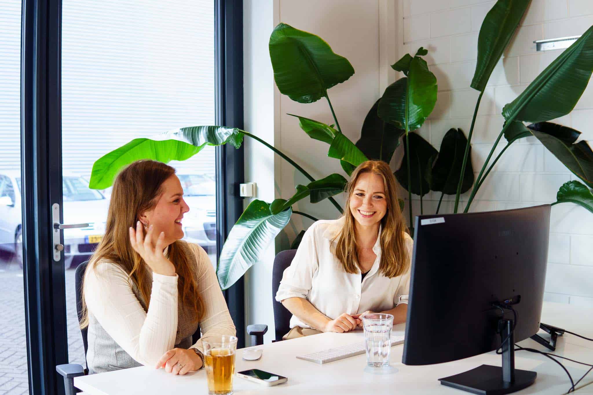 BURO210 Fullservice Marketingbureau Twee vrolijke collega's, vrouwen, lachen en werken samen achter een computer op kantoor met planten en drankjes op de tafel.