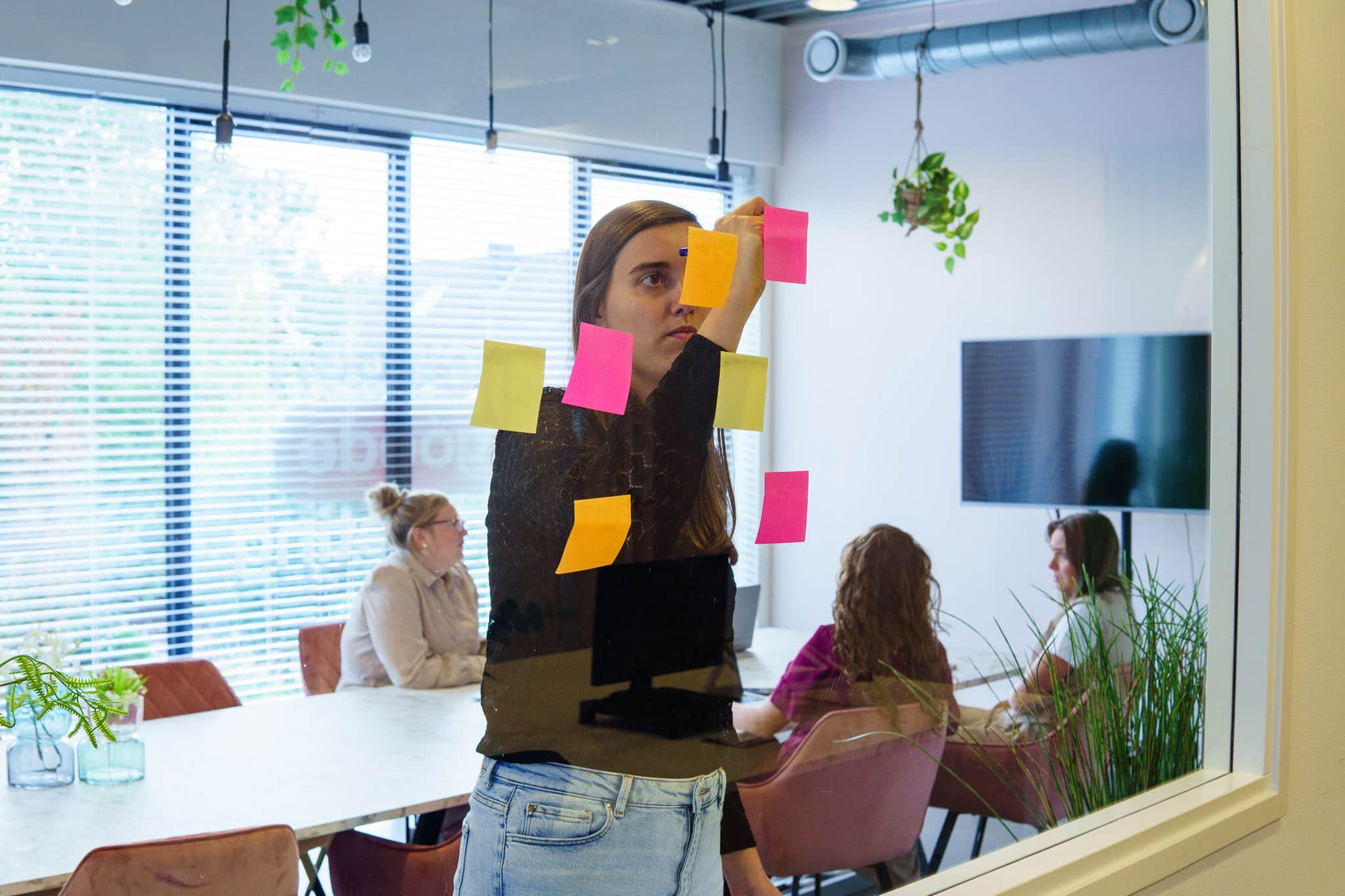 Een vrouw plakt gekleurde post-its op een glazen wand tijdens een brainstorm, terwijl collega’s aan een tafel in de achtergrond in gesprek zijn.
