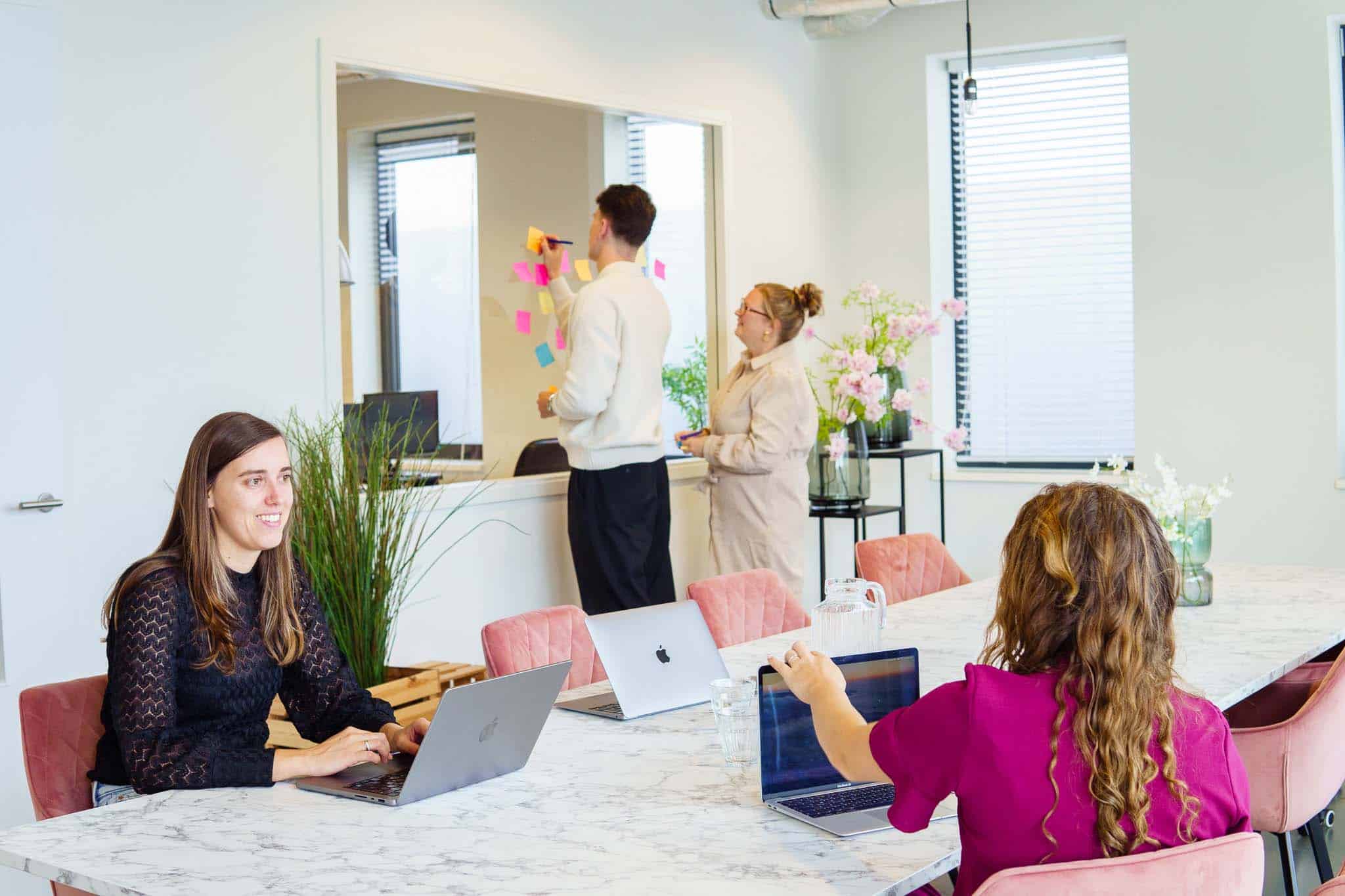 Teamleden werken aan laptops aan een grote tafel terwijl twee anderen post-its op een glazen wand plaatsen tijdens een brainstormsessie.