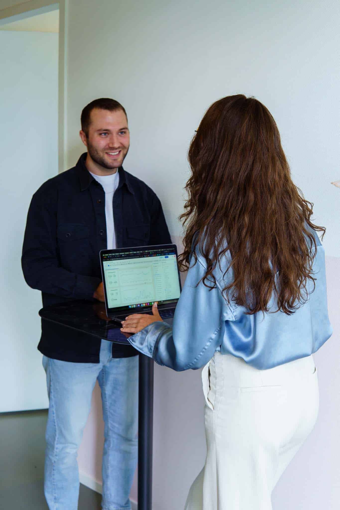 BURO210 Fullservice Marketingbureau Twee collega's bespreken data op een laptop in een modern kantoor. De man lacht en de vrouw laat grafieken en statistieken zien voor een zakelijke analyse.