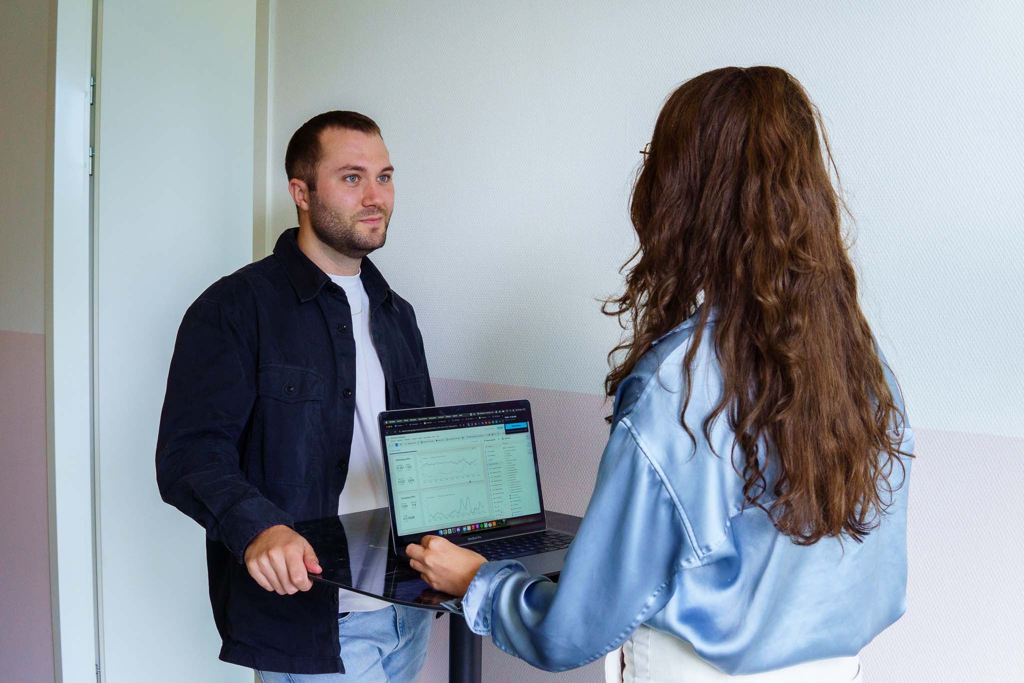BURO210 Fullservice Marketingbureau Twee jonge collega's bespreken data op een laptop in een lichte kantooromgeving, waarbij de vrouw de laptop vasthoudt en de man ernaar kijkt. Focus op samenwerking en datanalyse.