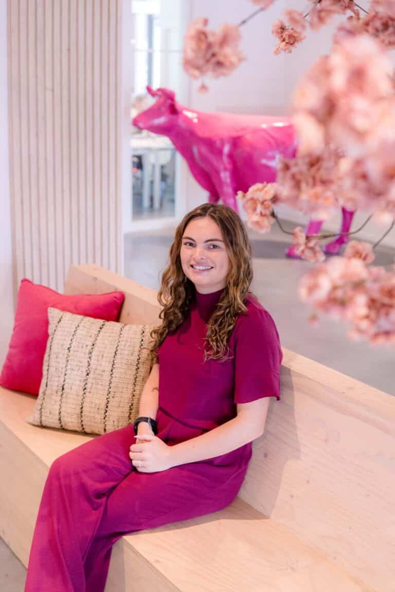 BURO210 Fullservice Marketingbureau Portret van een lachende vrouw in een bordeauxrode jumpsuit, zittend op een houten bank met kussens, met een roze koe sculptuur en kersenbloesem op de achtergrond.