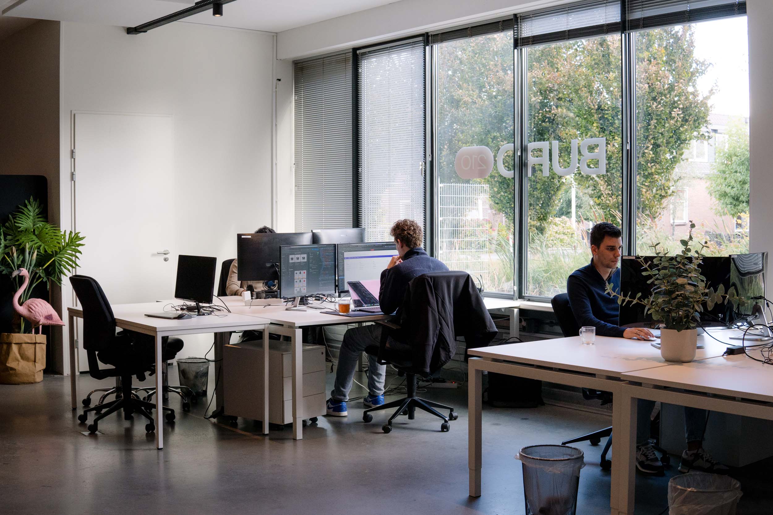 BURO210 Fullservice Marketingbureau Moderne kantoorruimte met mensen die aan witte bureaus werken, computers en grote ramen die uitzicht bieden op een groene omgeving. Productieve en lichte werkplek.