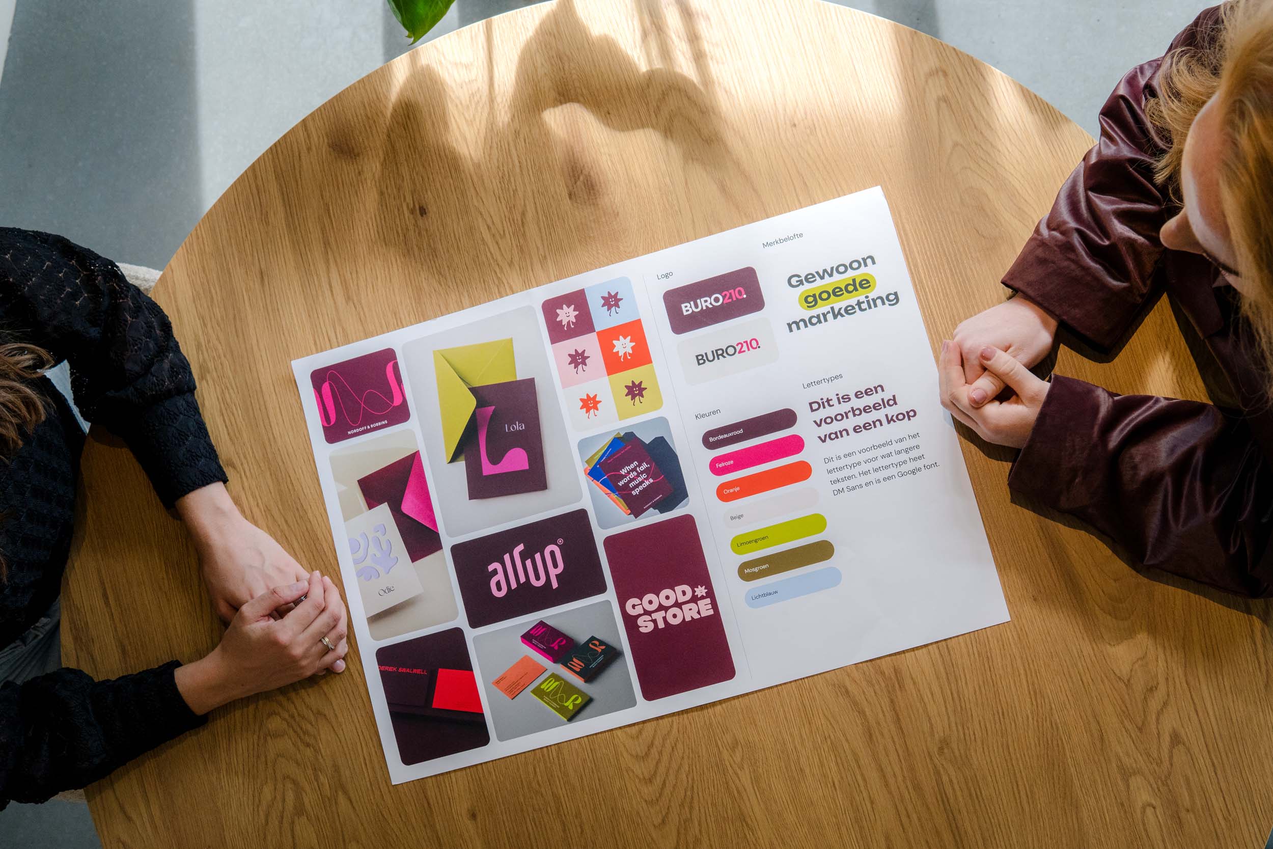 BURO210 Fullservice Marketingbureau Twee personen bekijken een marketingoverzicht met logo's, kleurenpaletten en lettertypes van BURO210, gericht op 'Gewoon goede marketing'.