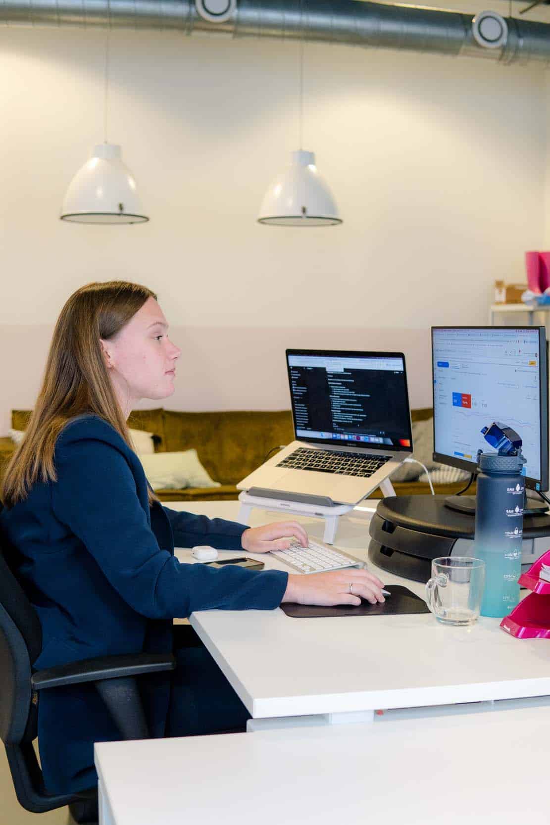 BURO210 Fullservice Marketingbureau Jonge vrouw werkt geconcentreerd aan haar bureau met laptop en monitor. Programmeur of data analist in een moderne kantooromgeving.