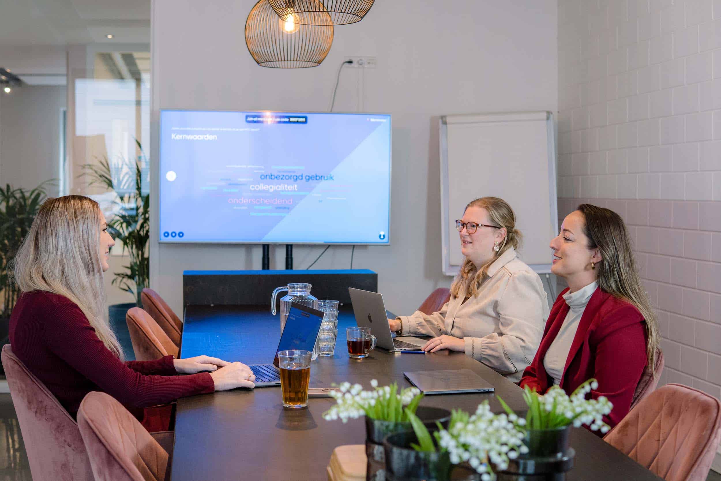 BURO210 Fullservice Marketingbureau Drie zakenvrouwen in een moderne vergaderruimte, werkend aan laptops met een presentatie over bedrijfskernwaarden op een groot scherm achter hen. Collega's samen aan het werk.