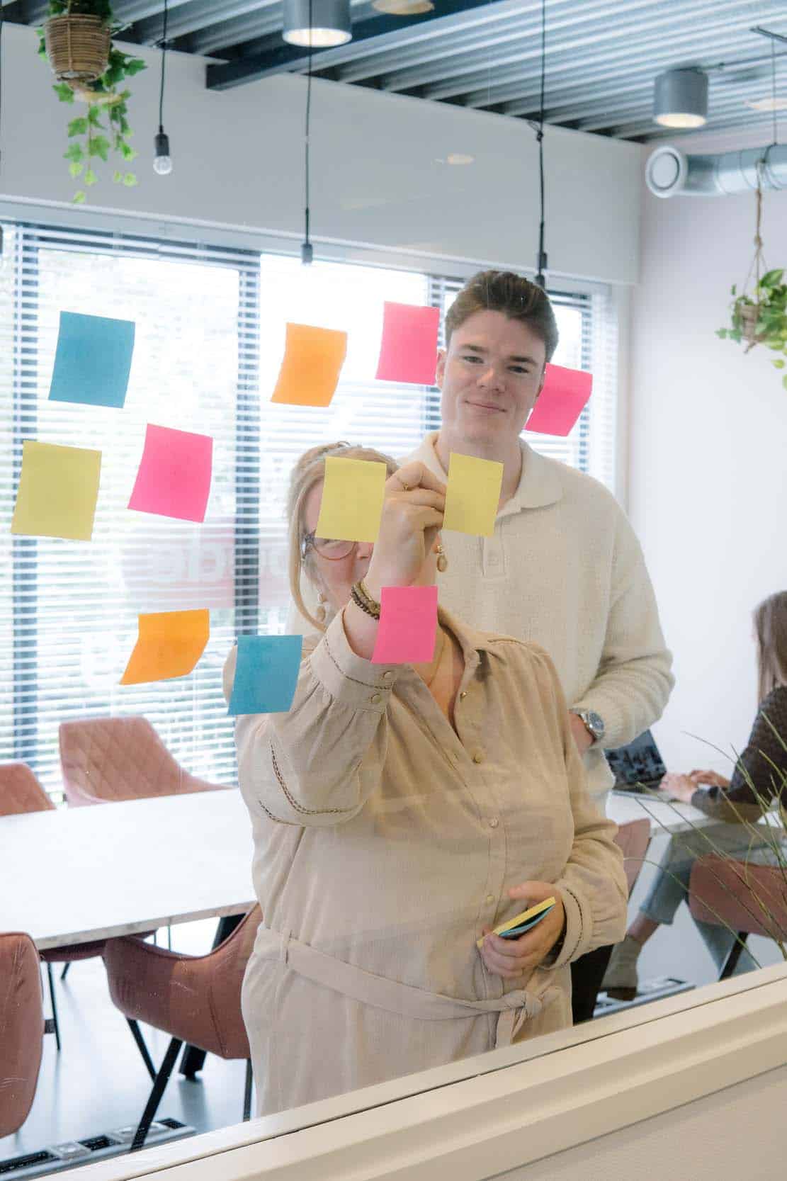 Twee personen plakken kleurrijke post-its op een glazen wand tijdens een brainstormsessie in een kantoorruimte.