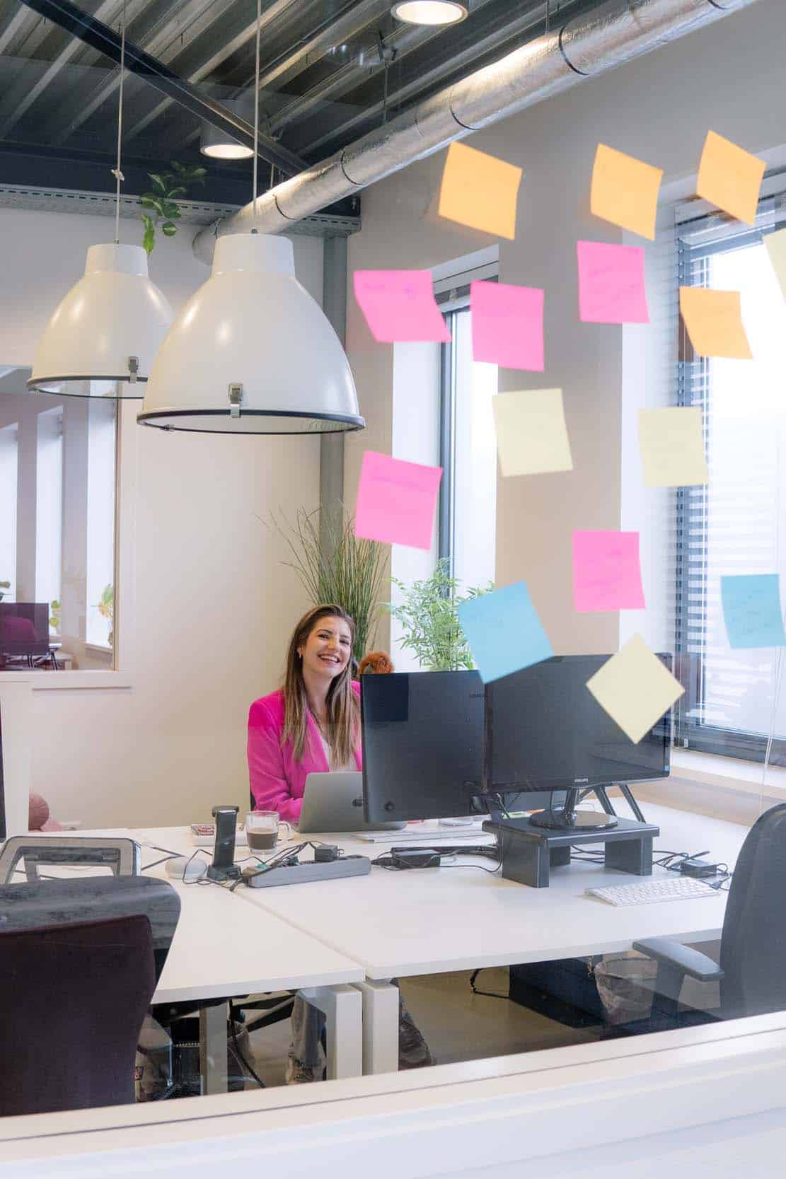 BURO210 Fullservice Marketingbureau Lachende jonge vrouw in een roze blazer werkt in een modern kantoor. Sticky notes op een glazen wand met computers, laptop, koffie en lampen.