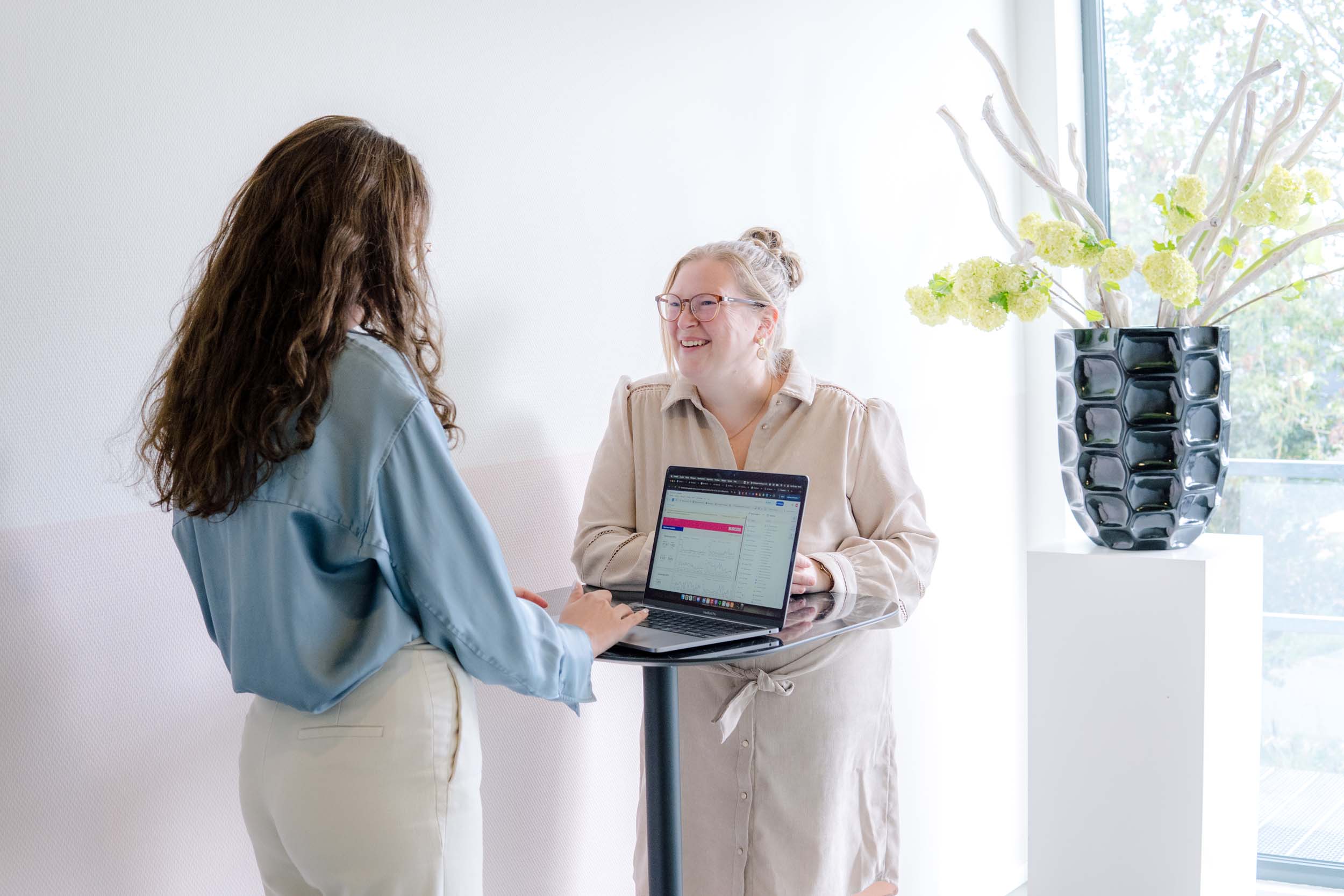 BURO210 Fullservice Marketingbureau Twee vrouwelijke collega's bespreken grafieken op een laptop in een moderne kantooromgeving, lachend en samenwerkend aan zakelijke projecten.