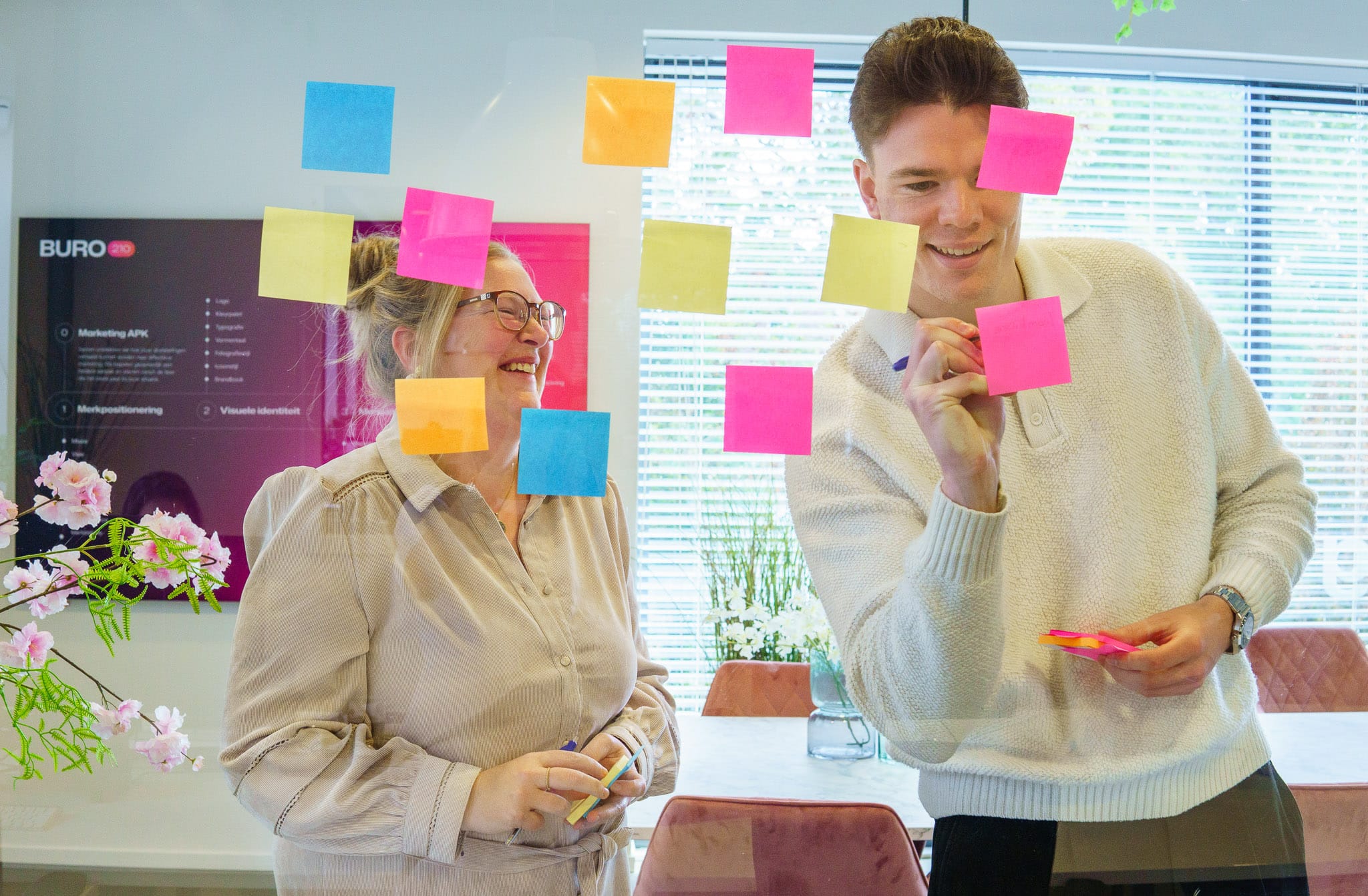 BURO210 Fullservice Marketingbureau Twee collega's, een man en een vrouw, brainstormen met kleurrijke post-its op een glazen wand in een modern kantoor, lachend en actief bezig met het genereren van ideeën.