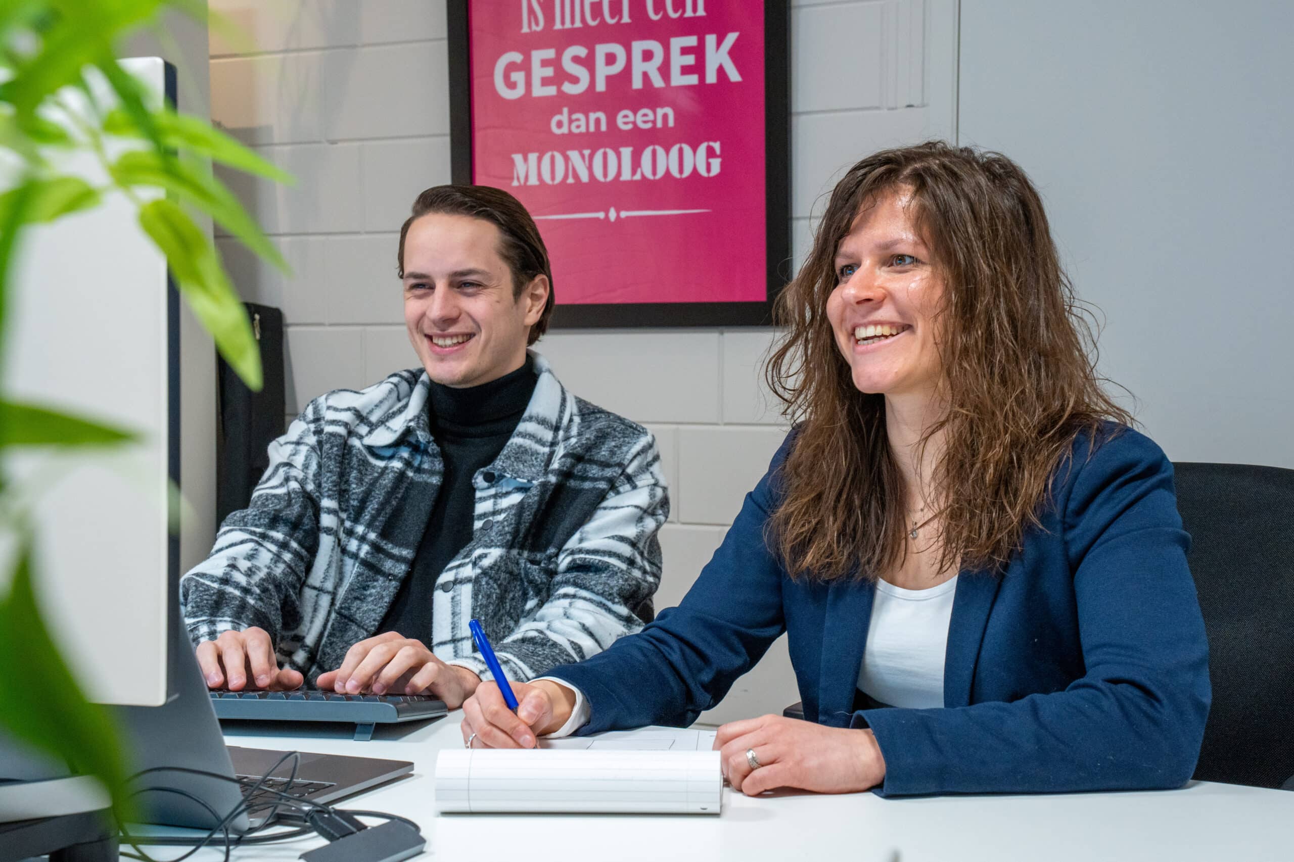 BURO210 Fullservice Marketingbureau Twee vrolijke collega's werken samen aan een nieuw project voor de visuele identiteit, een man typt op de computer en een vrouw maakt aantekeningen met een pen.