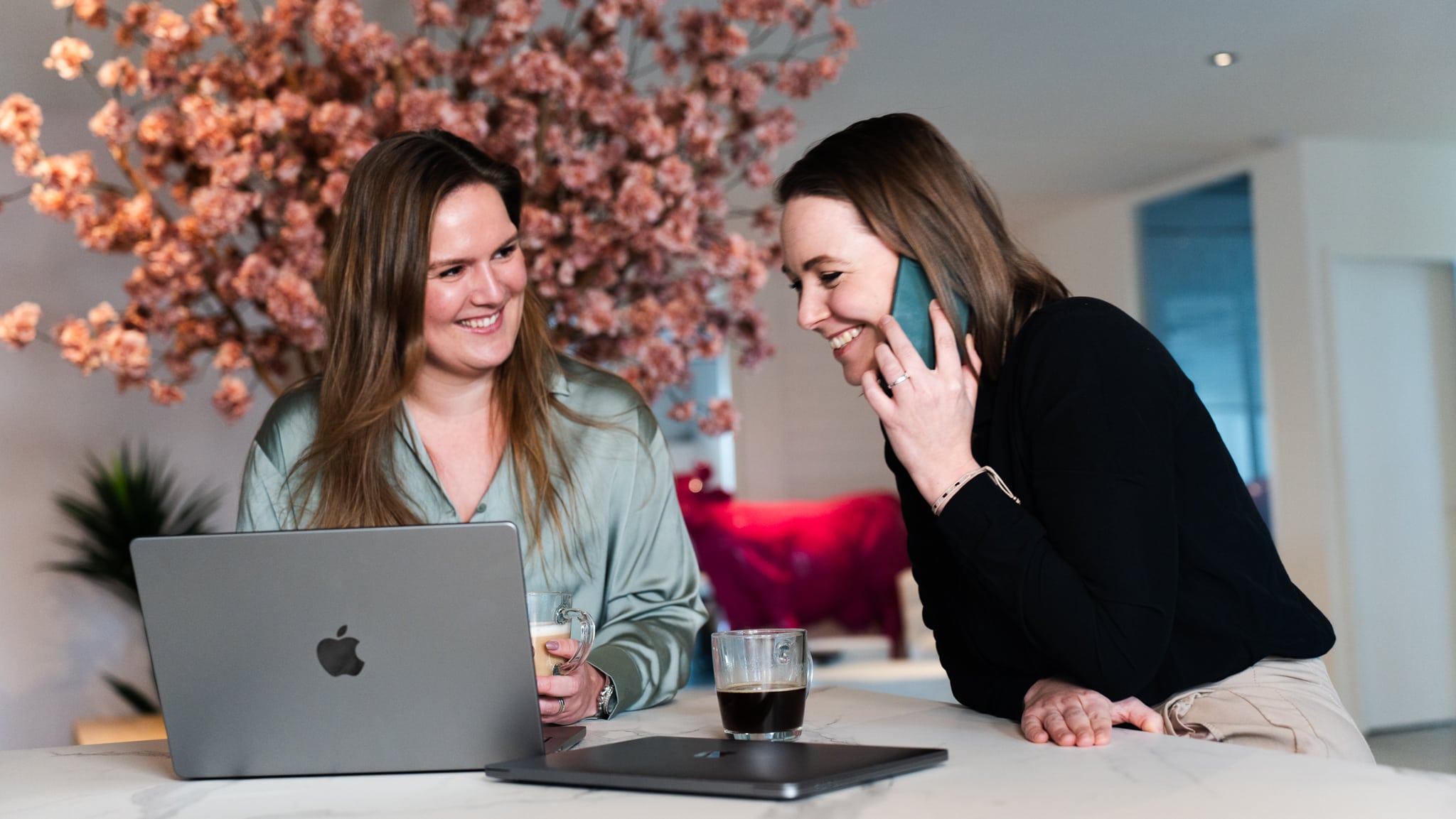 BURO210 Fullservice Marketingbureau Twee lachende vrouwen werken samen aan een laptop en bespreken de visuele identiteit van een bedrijf aan de telefoon. Marketing en branding concept.