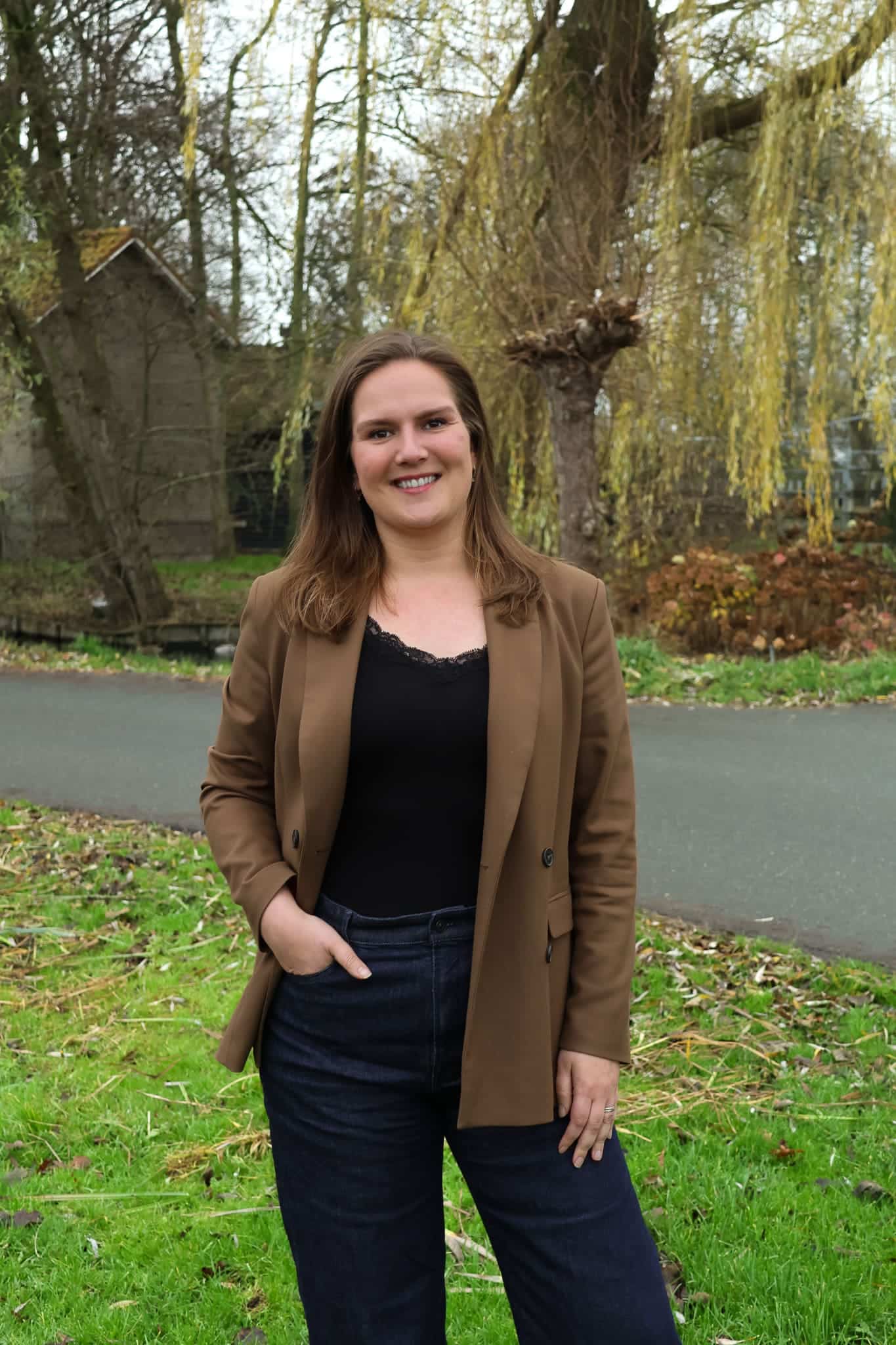 BURO210 Fullservice Marketingbureau Portret van een lachende vrouw met bruin haar en een bruine blazer in een groene omgeving met bomen op de achtergrond.