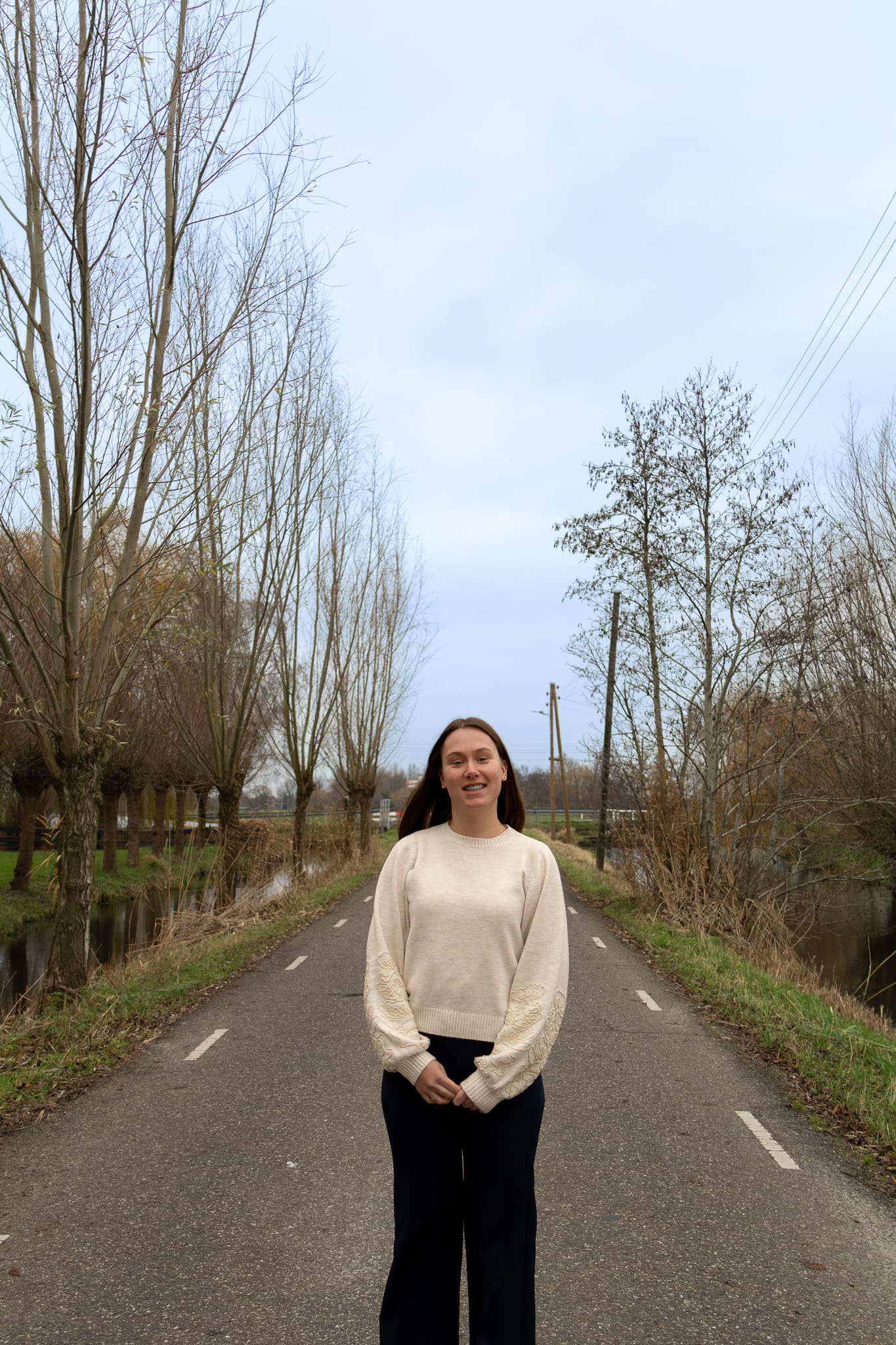 BURO210 Fullservice Marketingbureau Portret van een jonge vrouw op een landweg in Nederland. De weg wordt omzoomd door bomen en sloten, typisch voor het Nederlandse landschap.