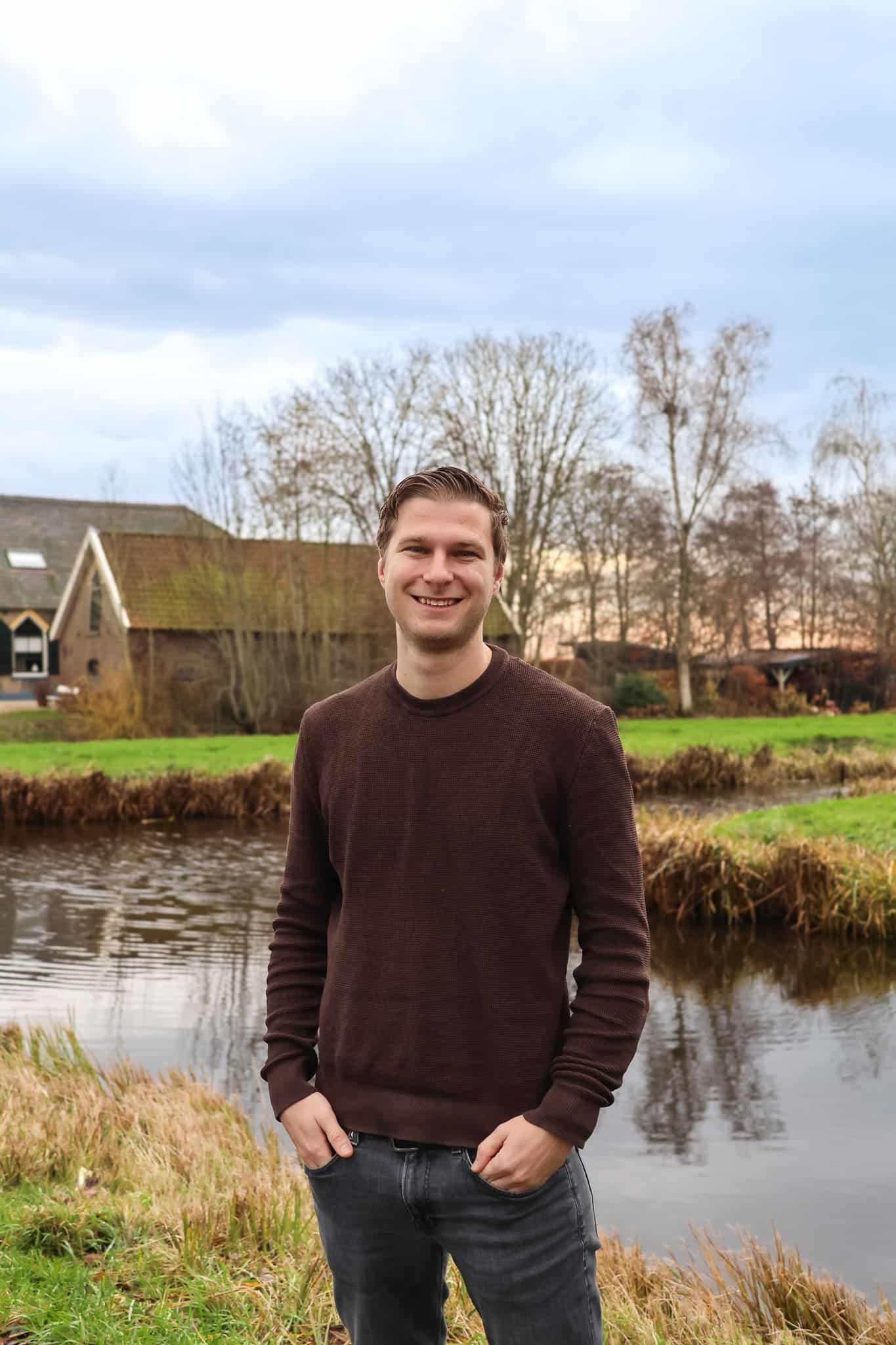 BURO210 Fullservice Marketingbureau Portret van een lachende man in bruine trui, staand bij een sloot met een traditionele boerderij op de achtergrond in een Nederlands landschap.